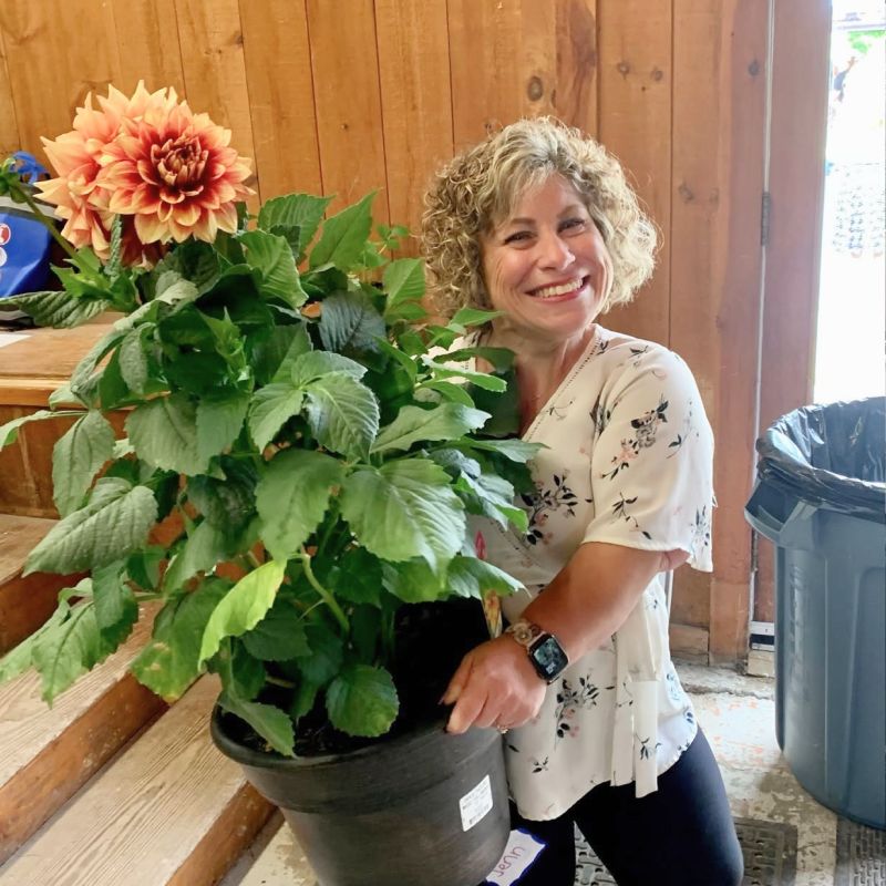 Smiling person in a floral blouse holding a large potted orange flower indoors