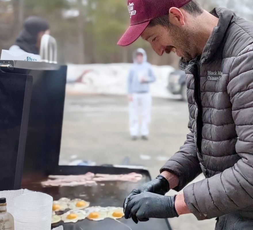 Man in red cap and gray jacket grilling food outdoors at a smoky street cookout