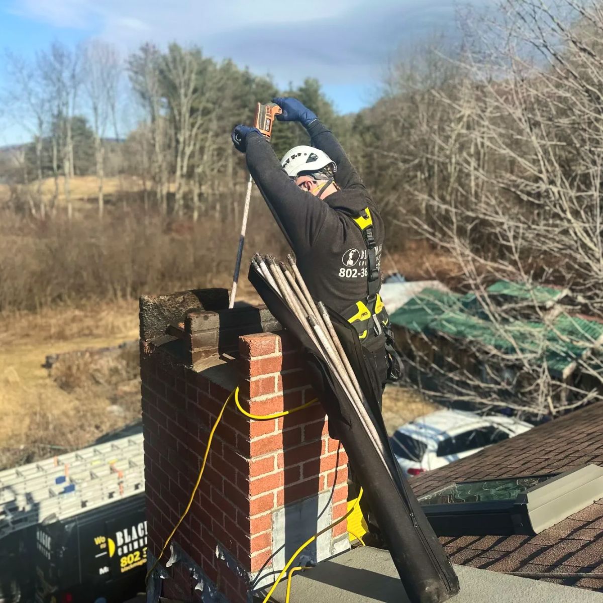 Worker on a rooftop chimney, hoisting a black pipe beside a red brick stack.