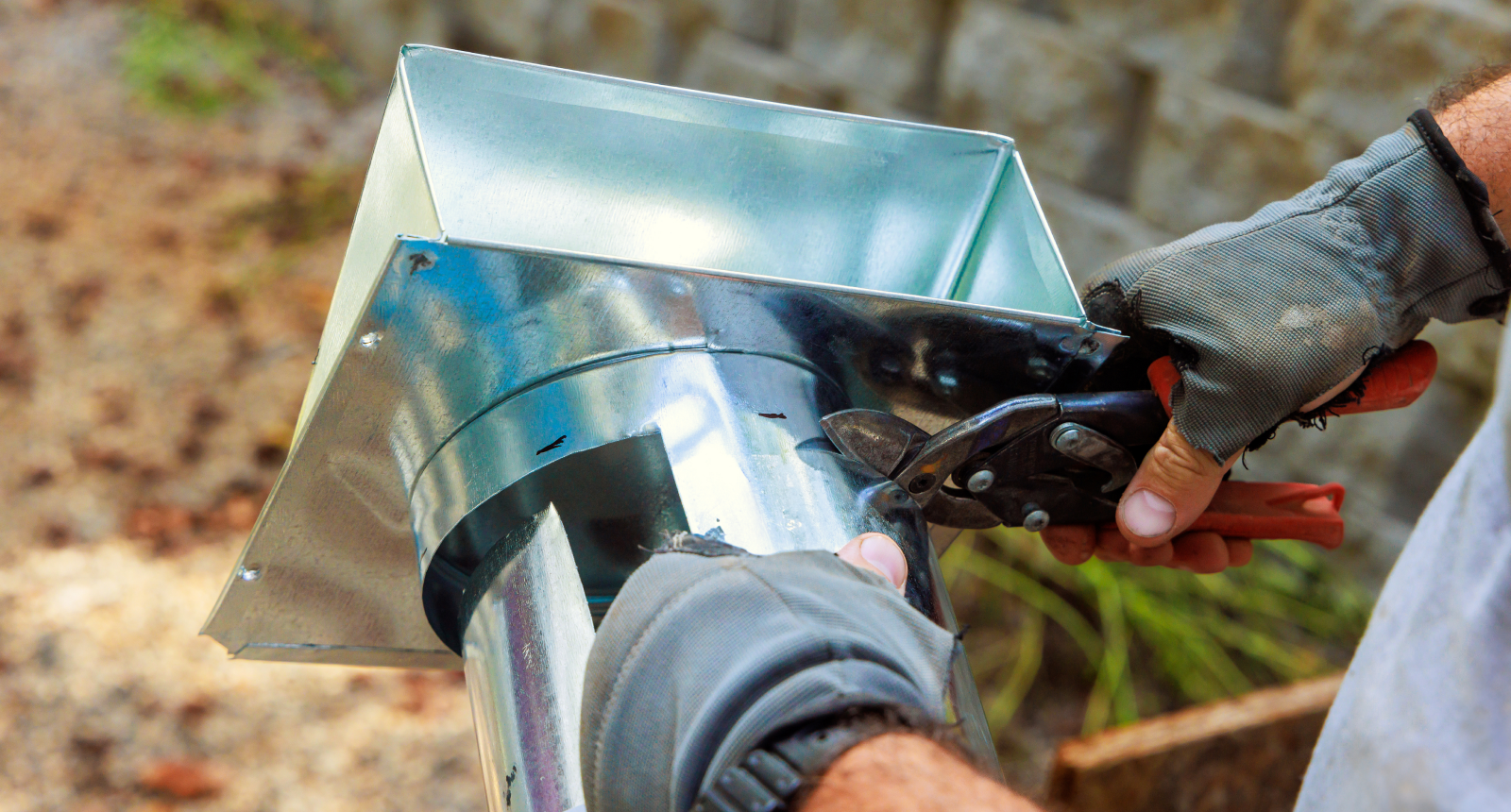 Hands in gray gloves cutting a shiny metal duct with snips outdoors