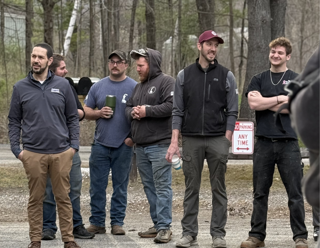Six people standing outdoors on a gravel path in a wooded area, wearing casual jackets and hoodies.