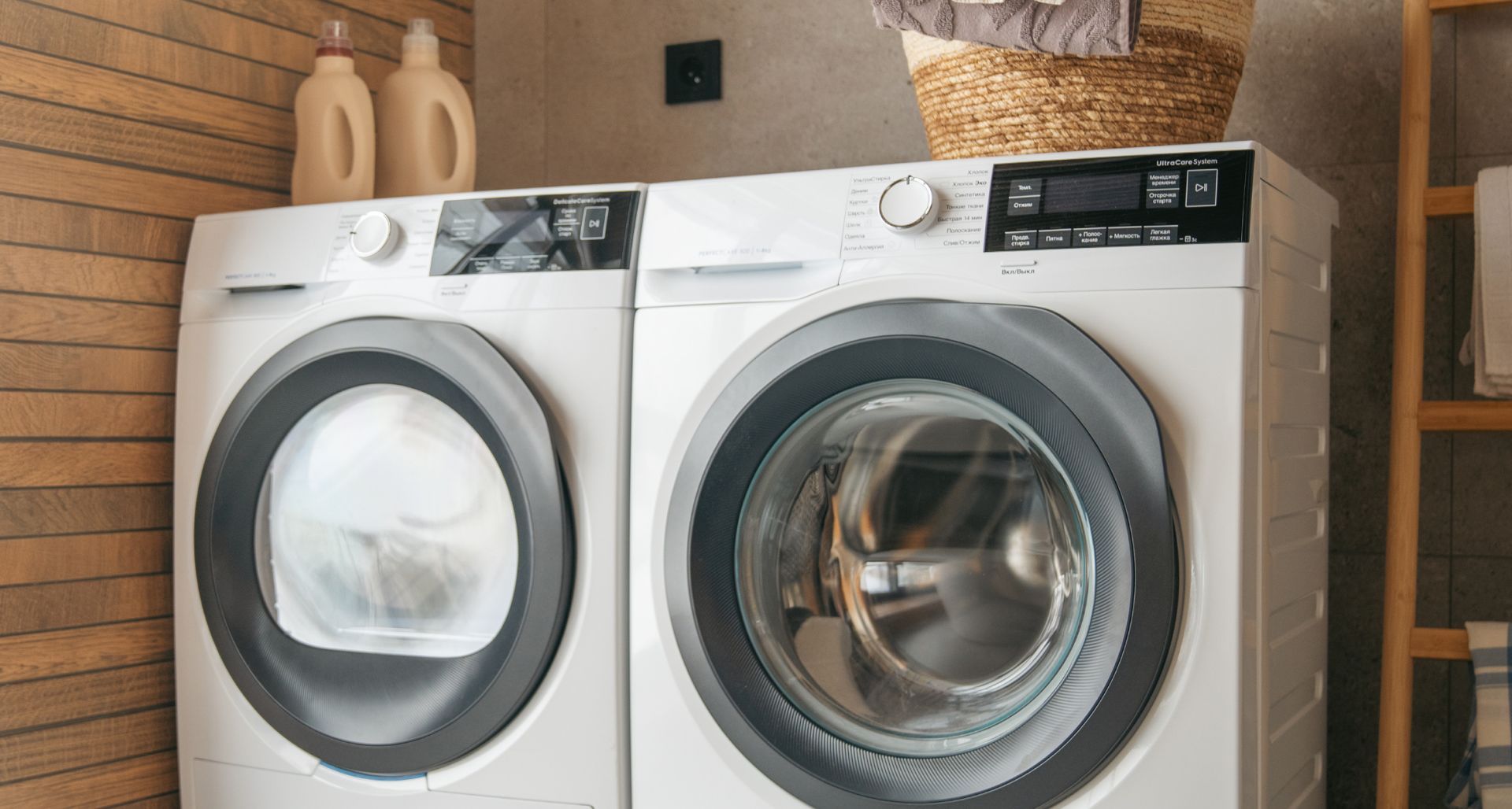 Two front-loading washers in a laundry room, one white and one silver, with bottles on a shelf above them