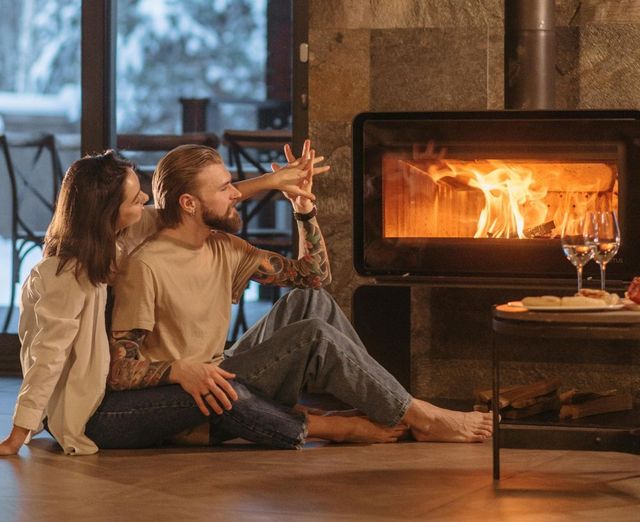 Couple sitting by a fireplace in a cozy living room, warming up near a roaring fire.