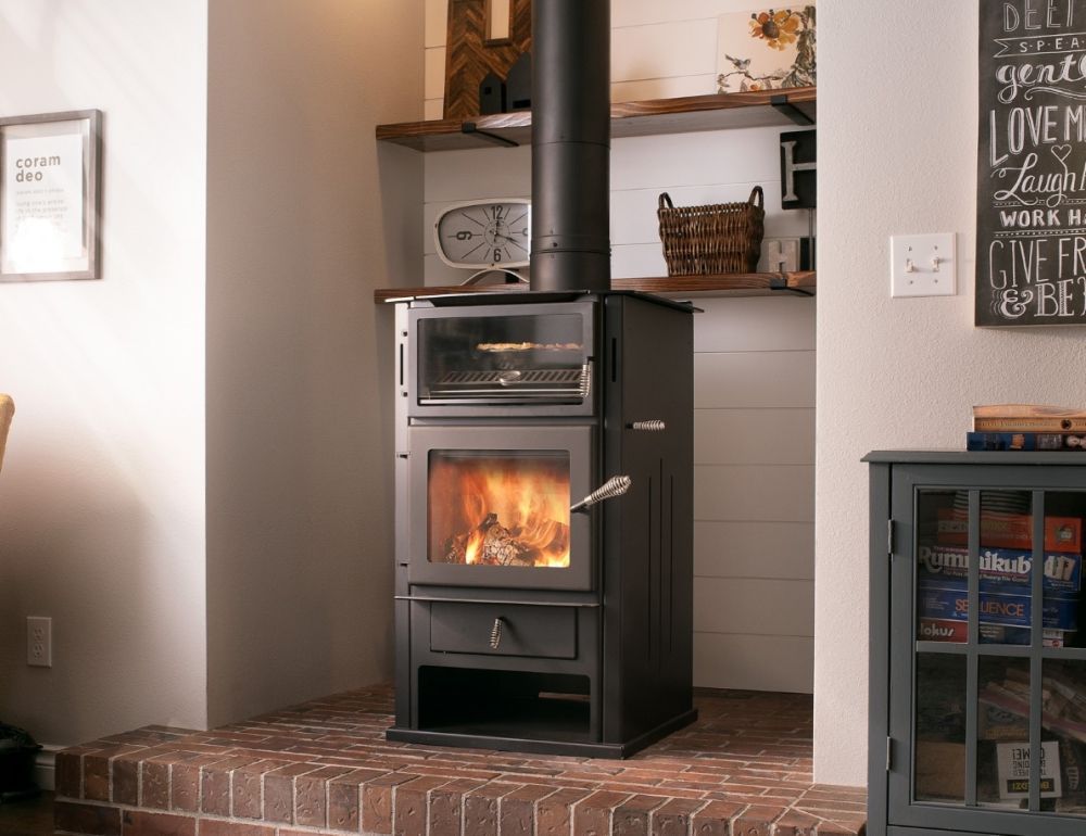 Black wood stove with visible fire in a brick hearth nook, beside wall decor and shelves.