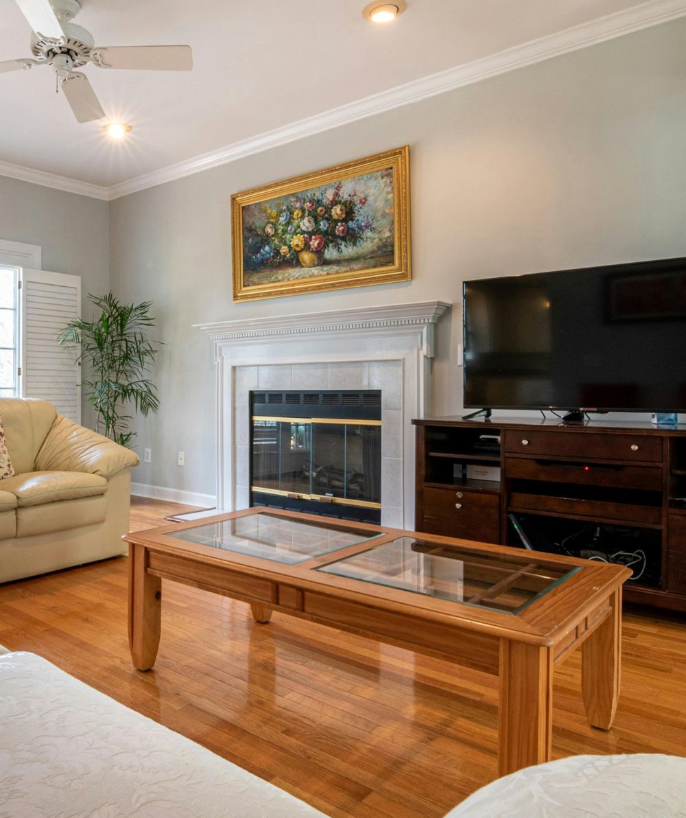 Bright living room with fireplace, TV, glass coffee table, tan sofa, and hardwood floors