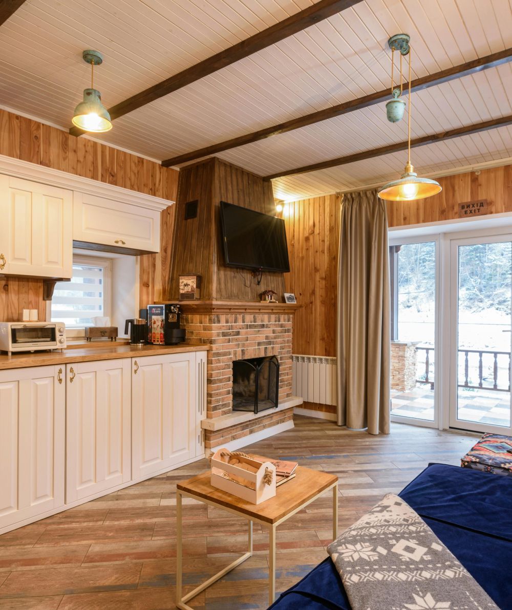 Cozy cabin living room with wood-paneled walls, fireplace, kitchenette, sofa, and snowy view through the window
