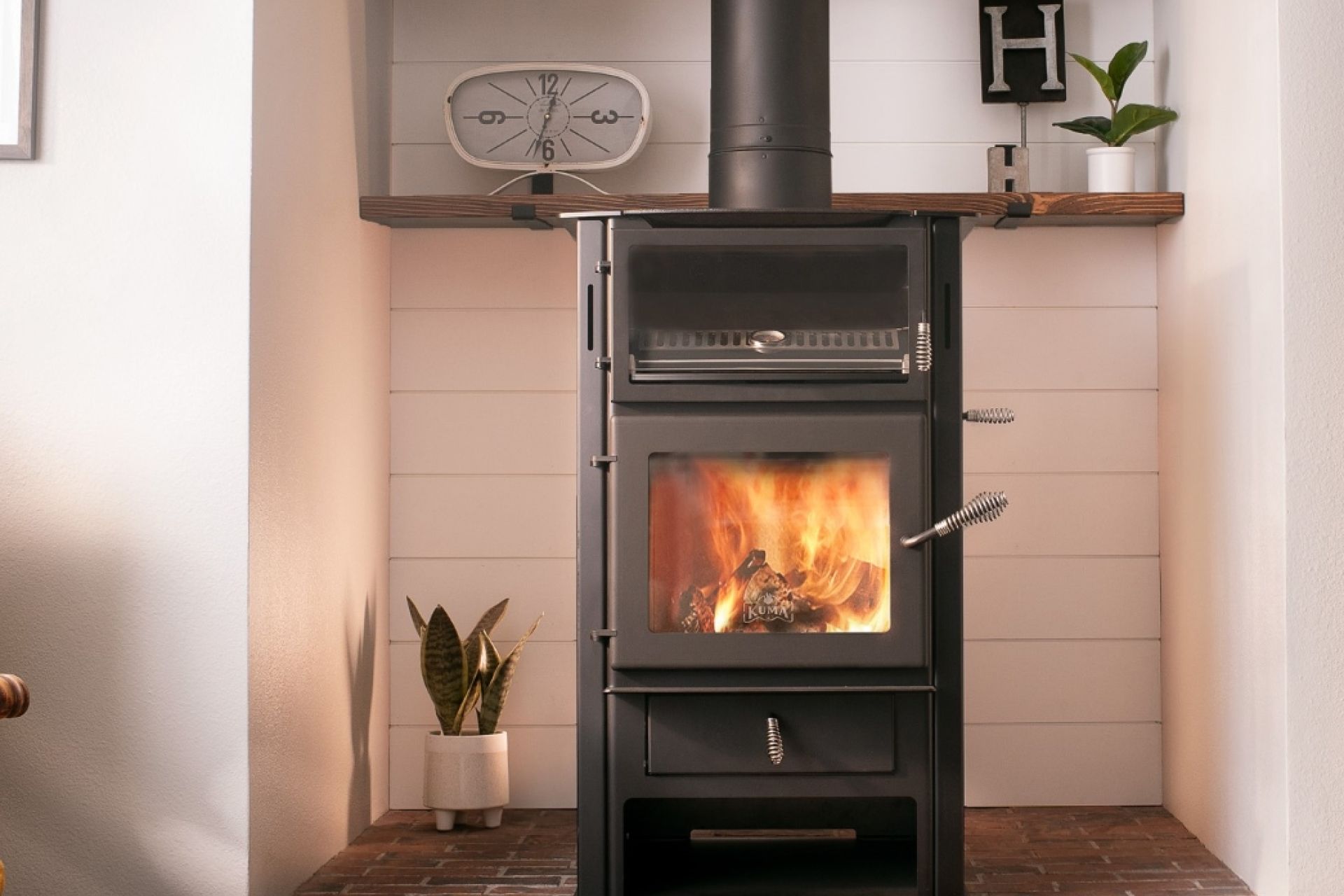 Black wood-burning stove with fire in a cozy room, flanked by plants and a shelf with decor.