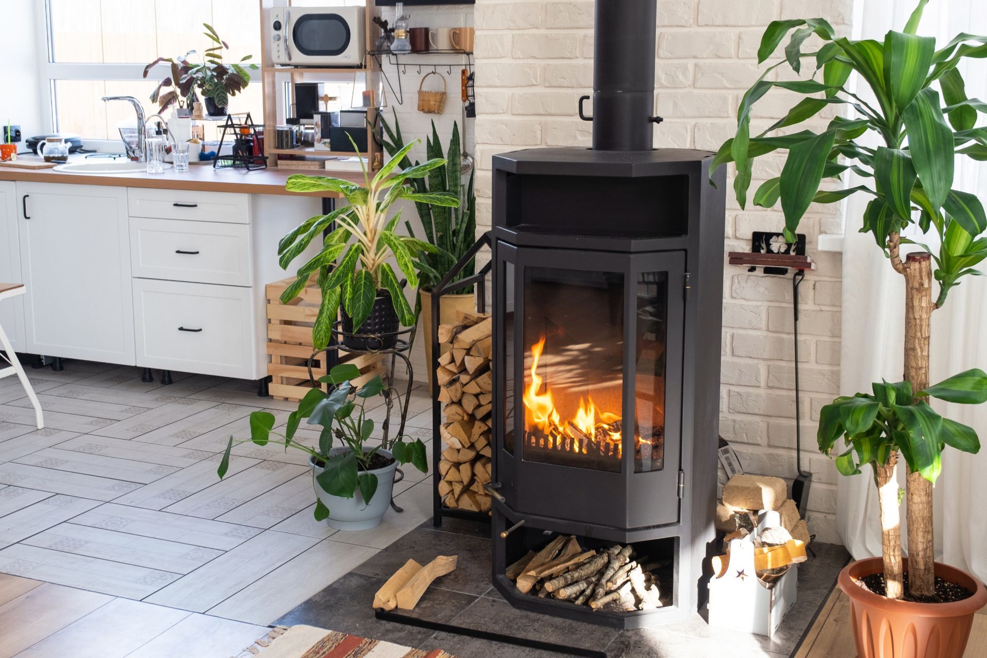 Cozy living room with a black wood stove, glowing fire, and potted plants beside a bright kitchen.