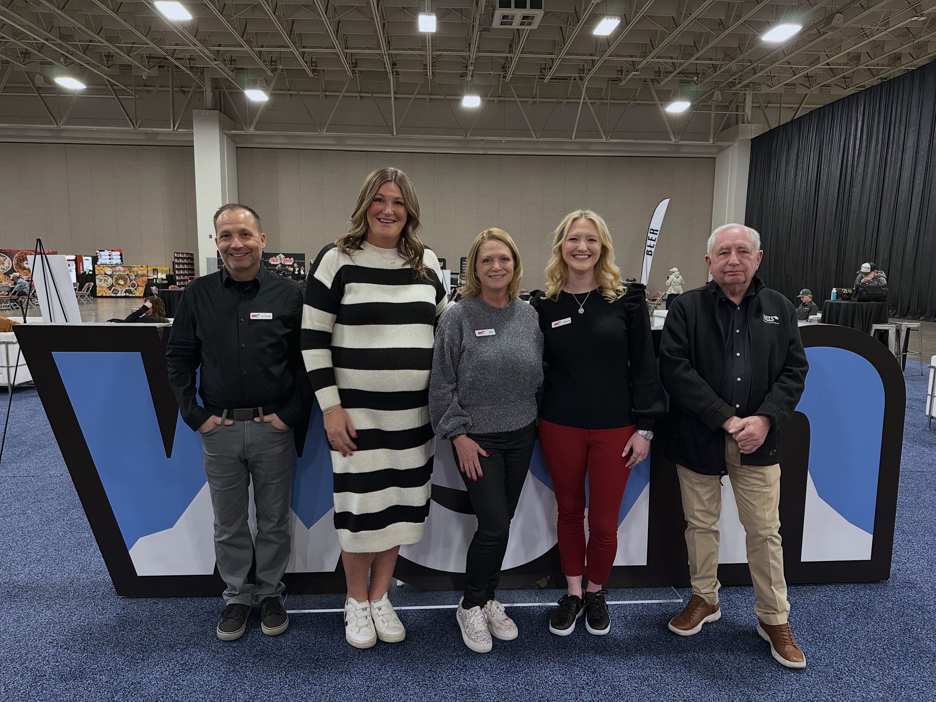 Five people posing together at an indoor event booth with a blue-and-black backdrop.