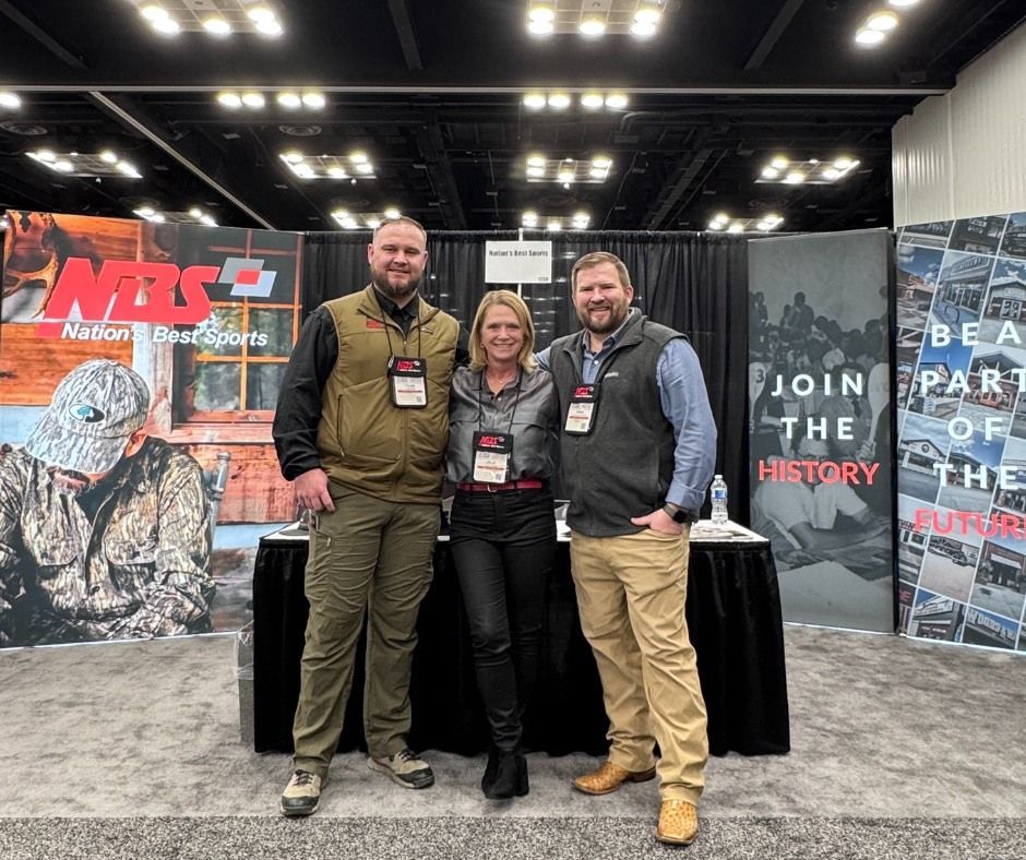Three people posing at a trade show booth with conference banners and a black table.