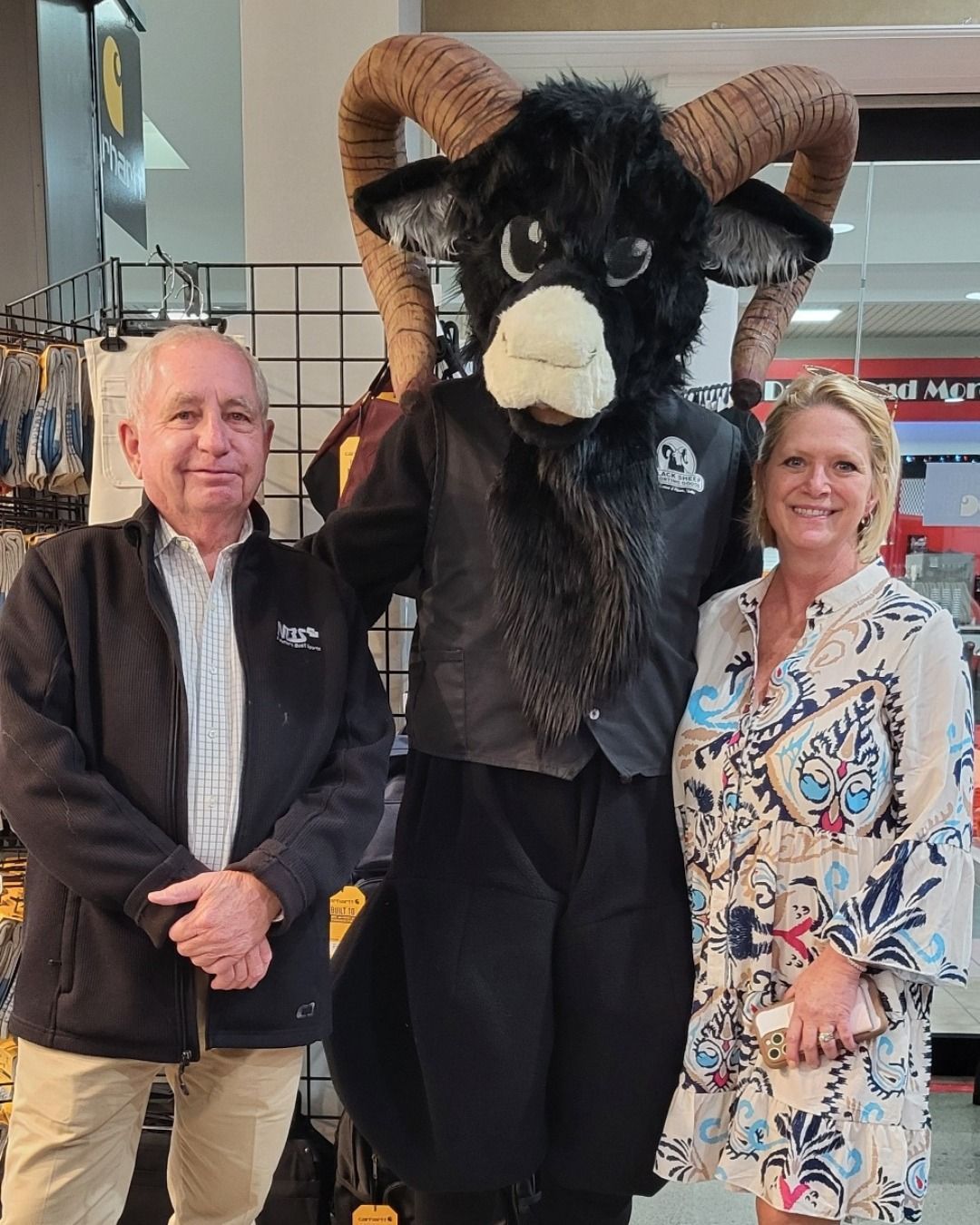 Two people standing beside a large black ram mascot with curled horns in a mall setting