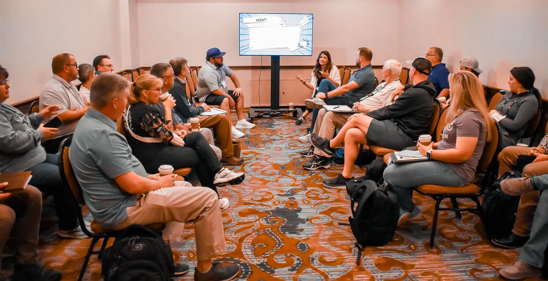 Group of people seated in a conference room, listening to a speaker near a screen.