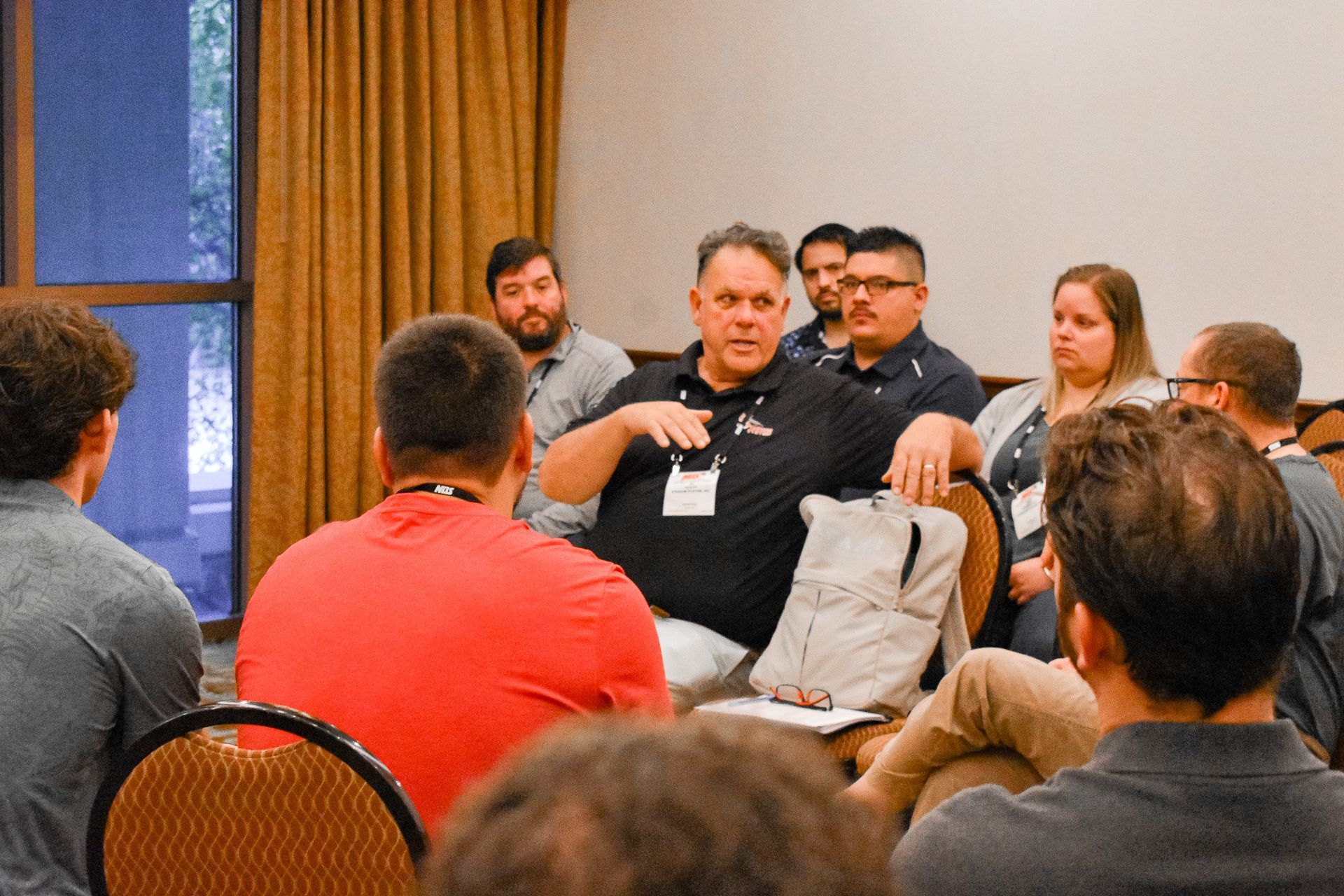 People seated in a discussion group in a room with a window and gold curtains.