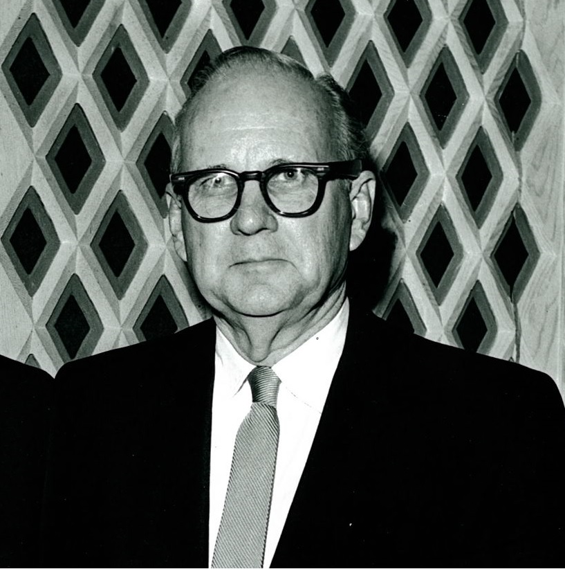 Black-and-white portrait of a man in glasses and a suit before a patterned backdrop