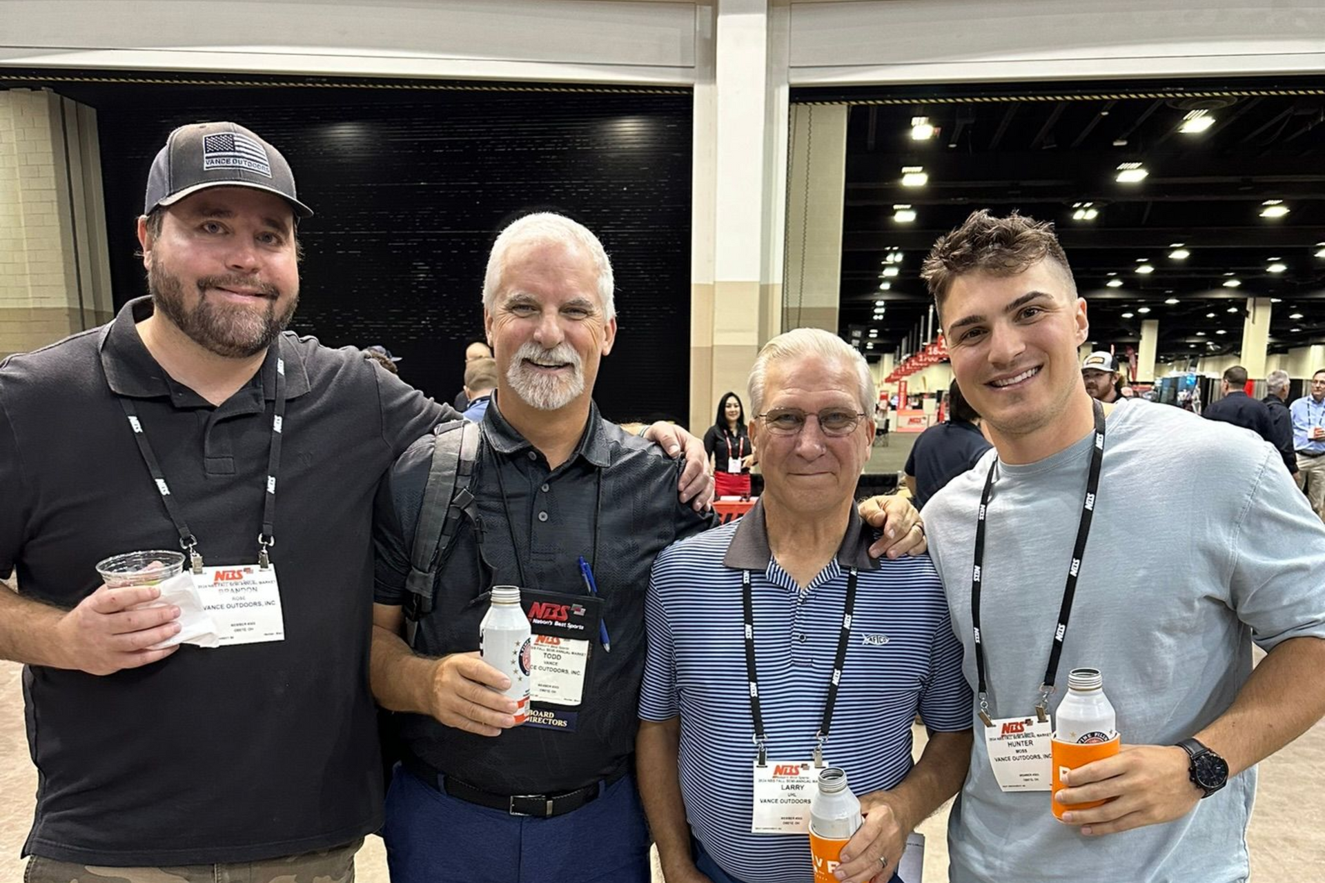 Four people posing at an indoor event, smiling and holding drinks and badges.