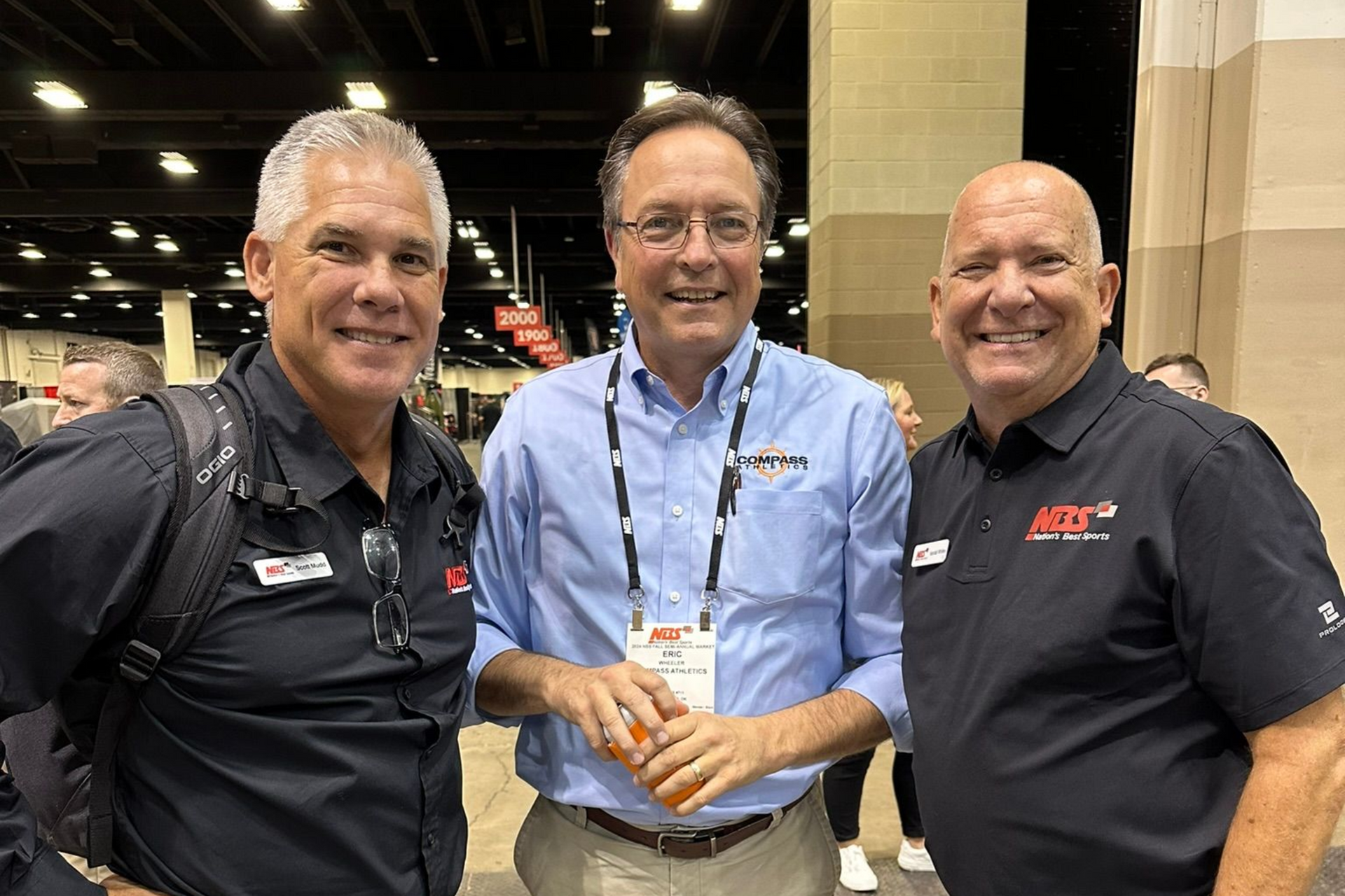 Three men smiling at an indoor event, wearing lanyards and business shirts, standing together.