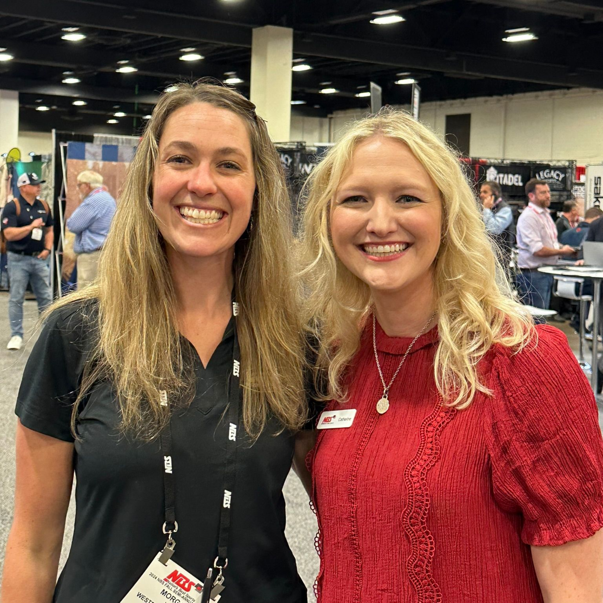 Two smiling women standing at a conference booth, one in black and one in red.