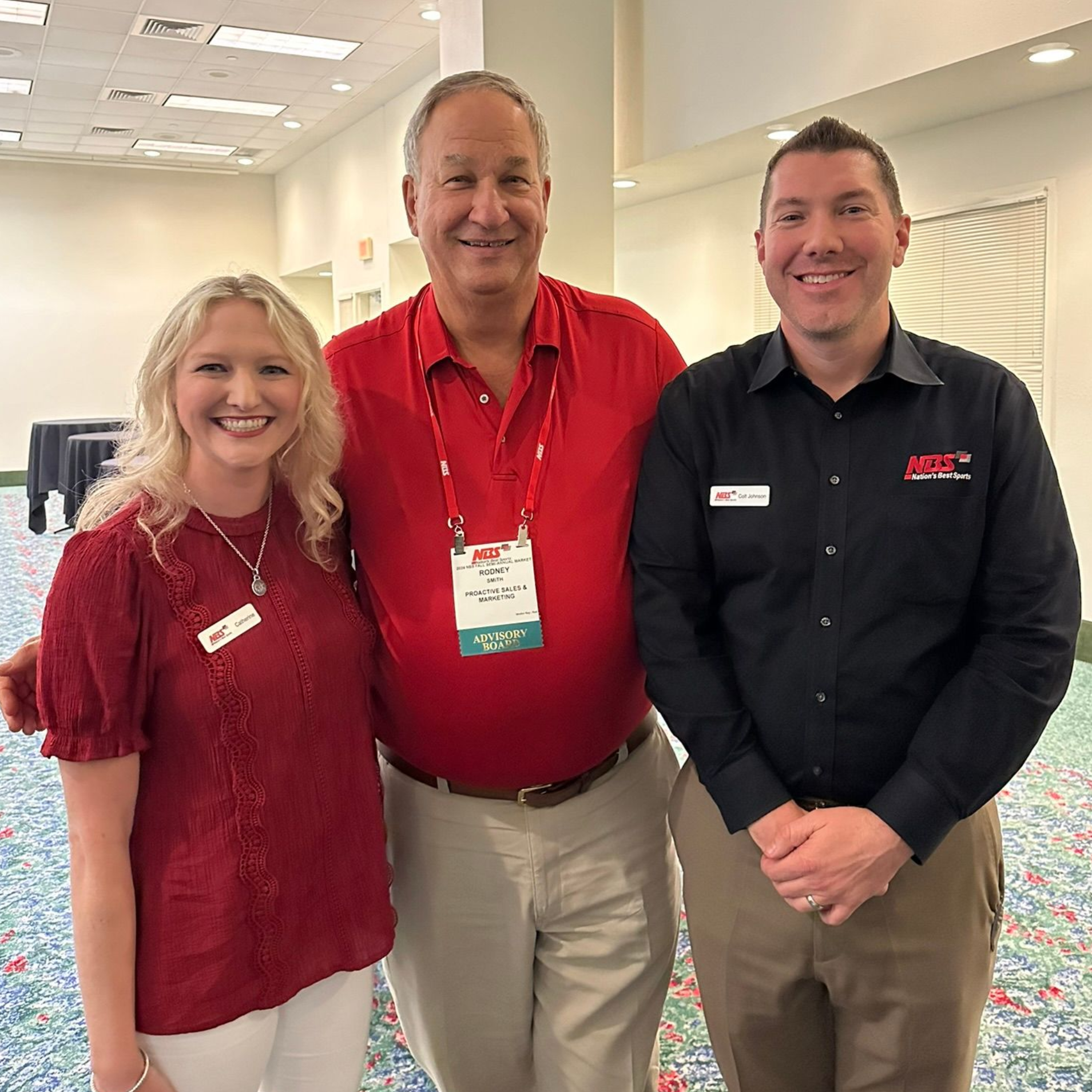 Three colleagues posing in a bright meeting room, smiling and wearing red and black company shirts.