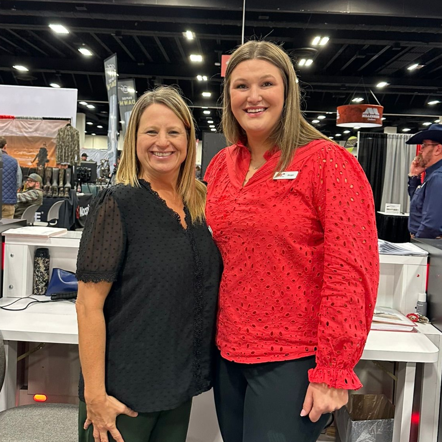 Two women smiling at an indoor trade show booth, wearing black and red tops.
