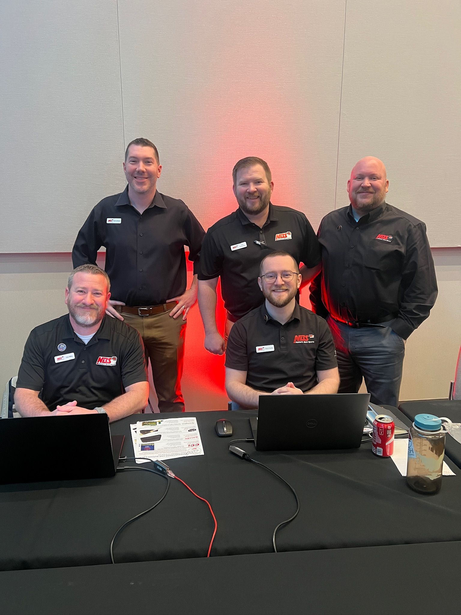 Five people in black shirts pose behind a table with laptops at an event booth