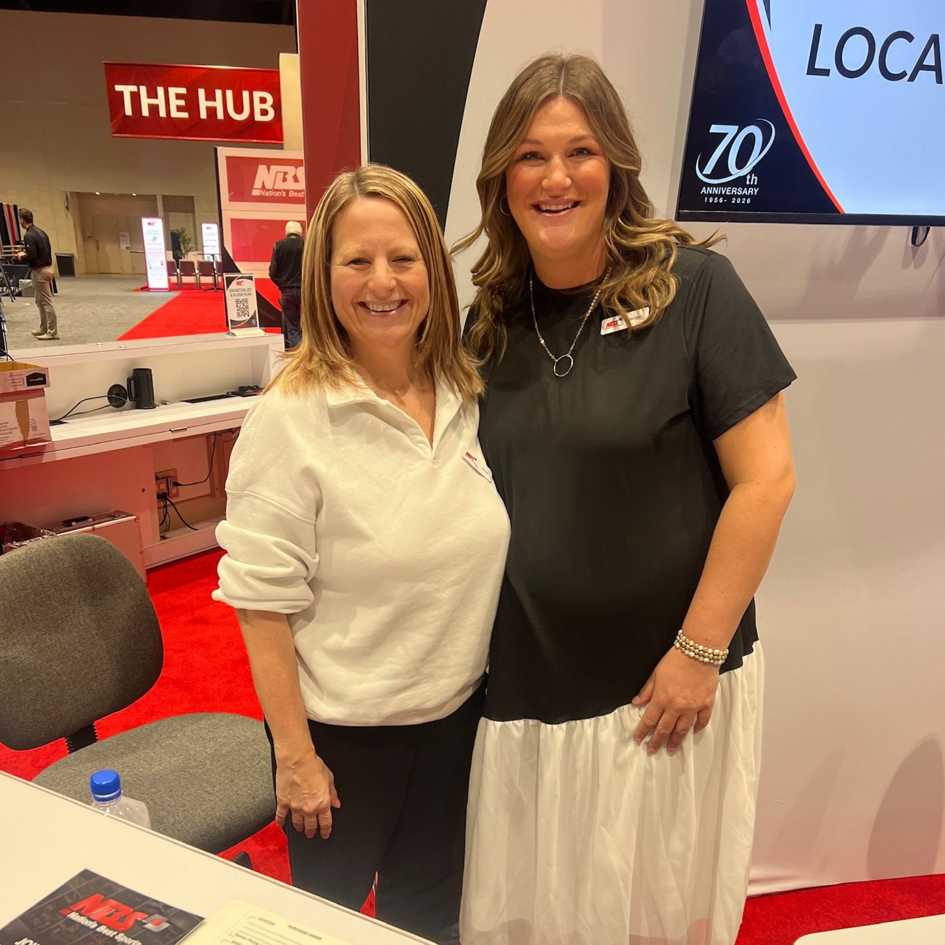 Two women smiling at a booth with red and white signage at an indoor event