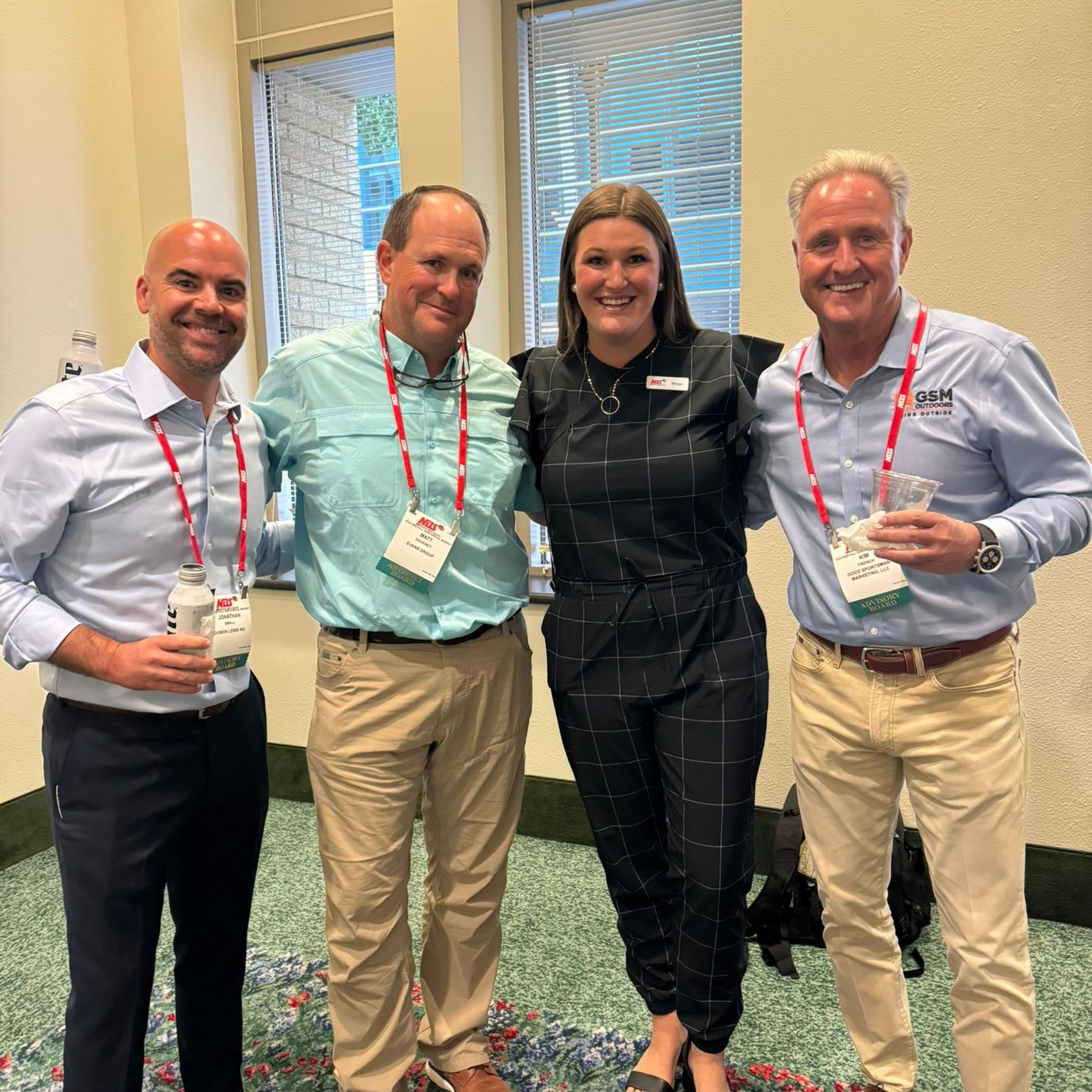 Four people posing indoors at a conference, wearing lanyards and holding drinks.