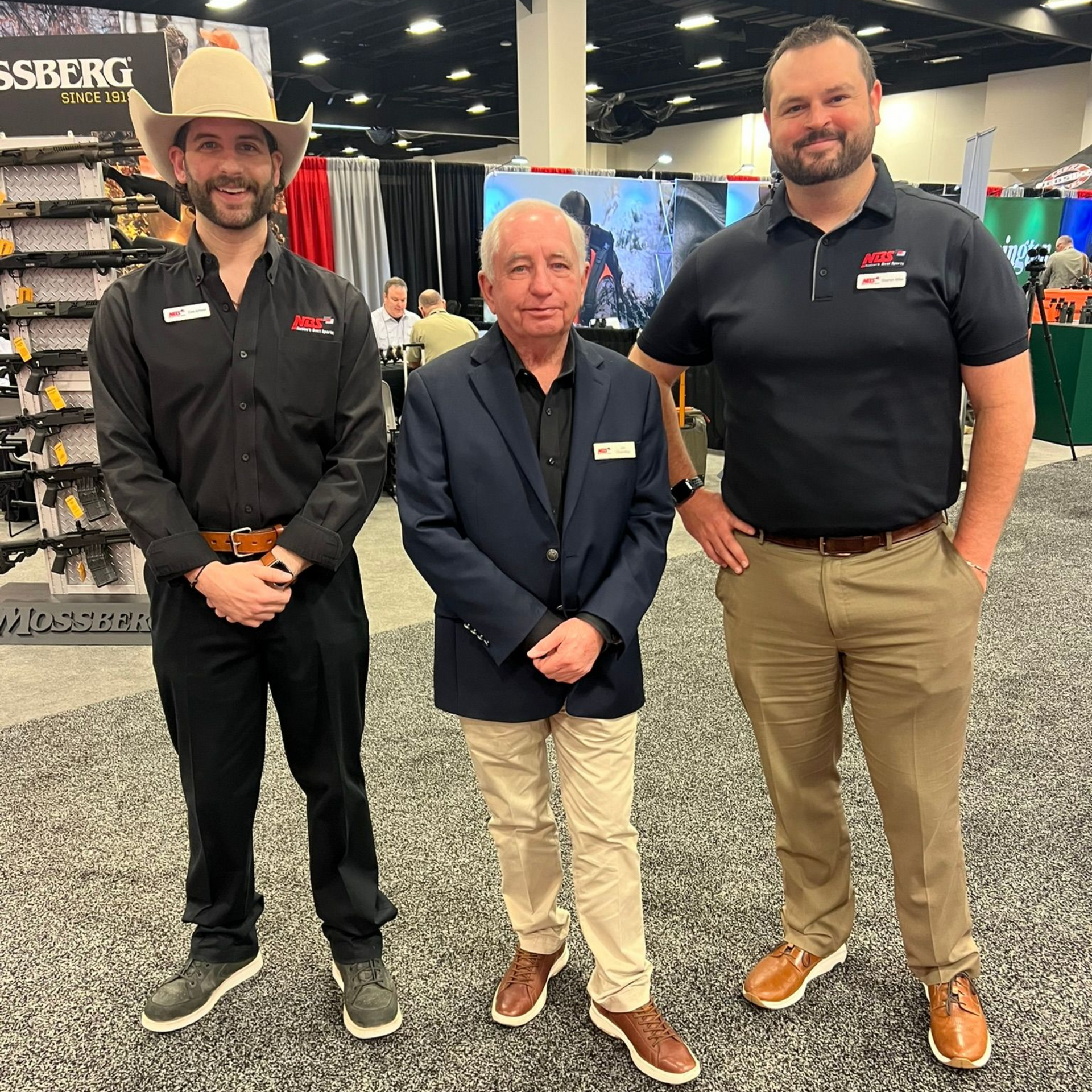Three men posing at a trade show booth, two in black shirts and one in a navy suit.