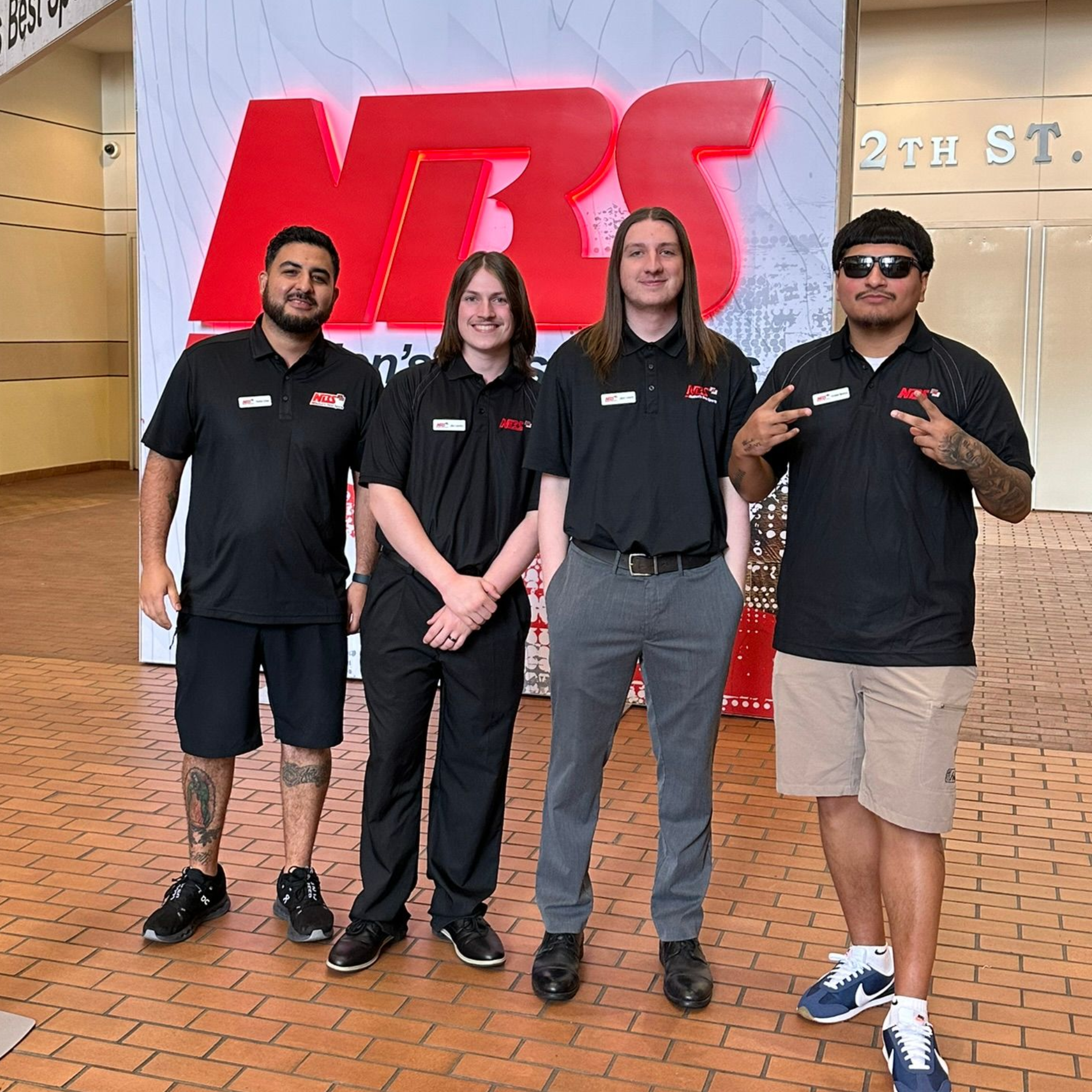 Four people in black shirts posing in front of a red NBS sign at 21st St.