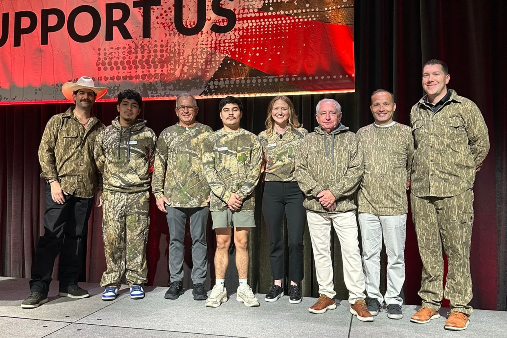 Eight people pose onstage before a red “WE SUPPORT THOSE WHO SUPPORT US” banner, some in camouflage uniforms.
