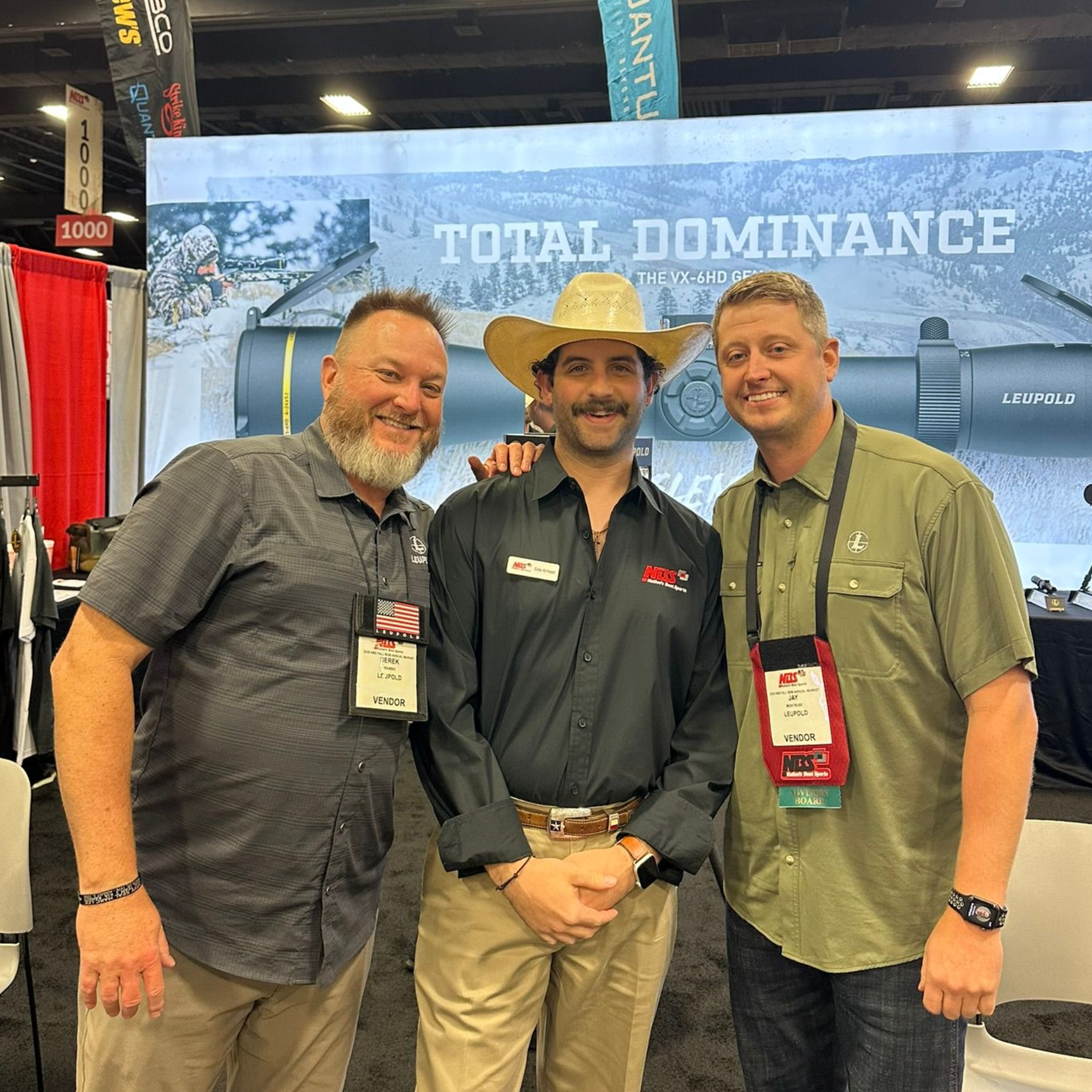 Three men posing together at a trade show booth with a “Total Dominance” banner in the background.