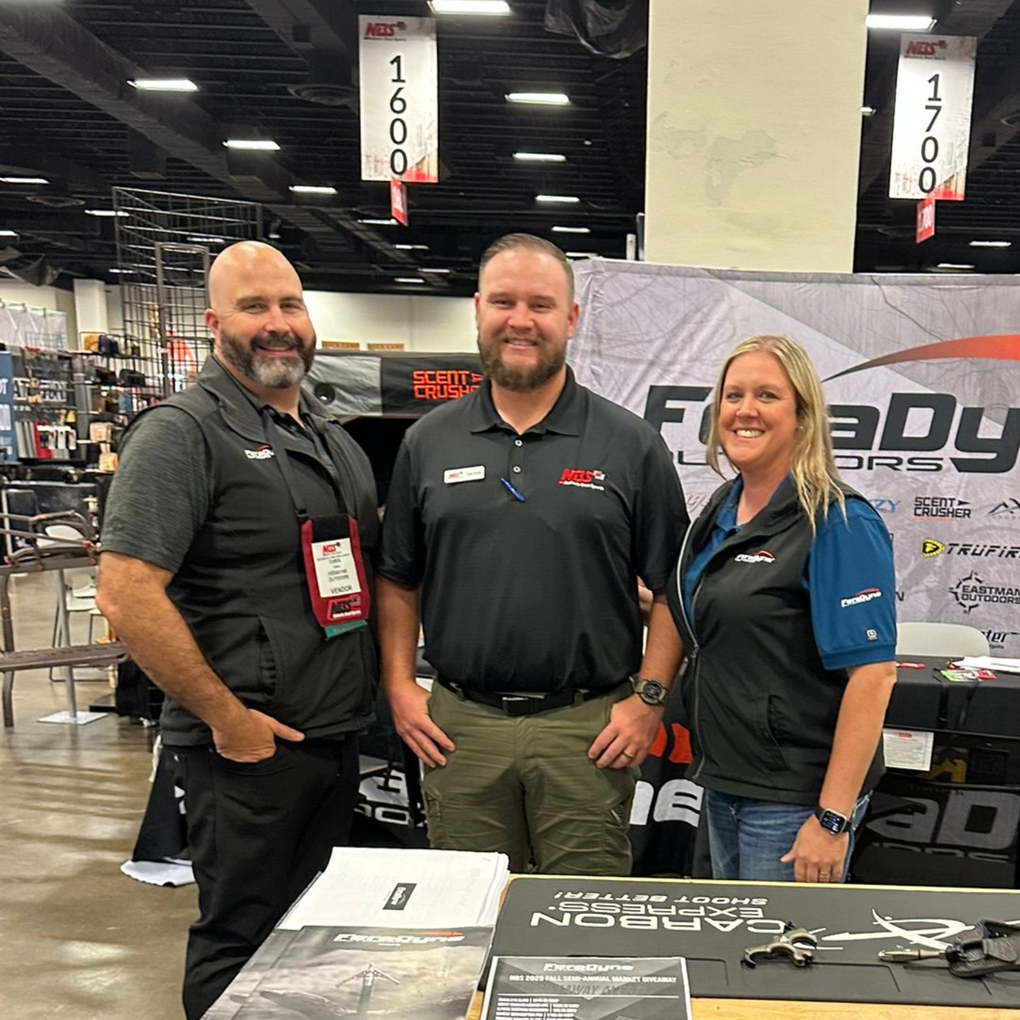 Three people posing at a trade show booth with a branded table display and banners behind them