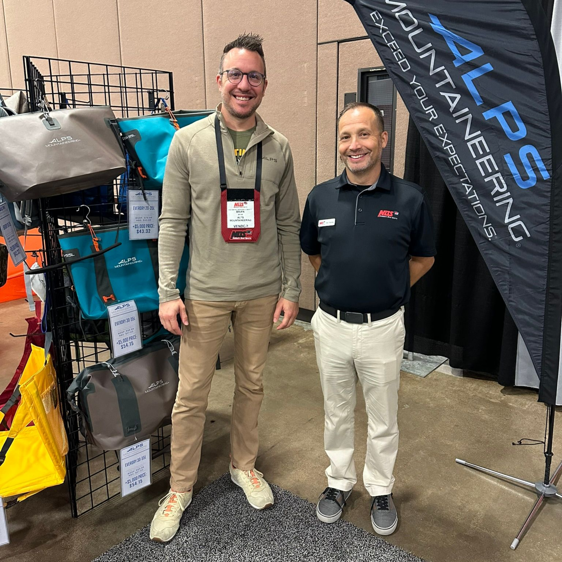Two men standing at a trade show booth with outdoor gear and a vertical ARS flag.