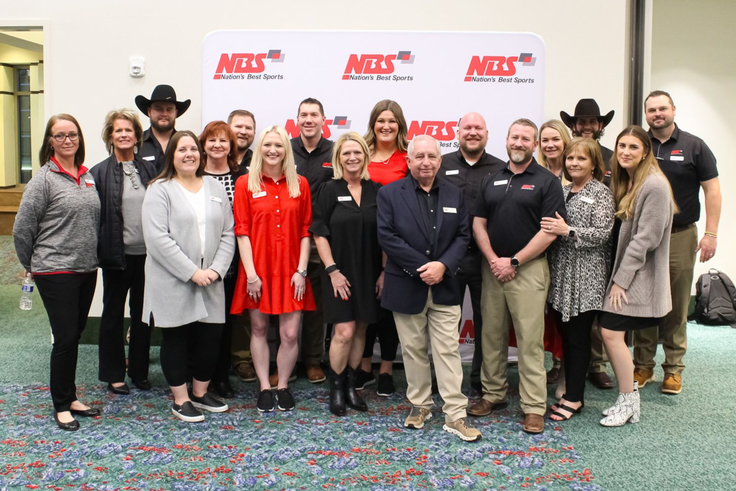 Group of people posing at an NBS event banner indoors, with one person in a bright red dress in the center.