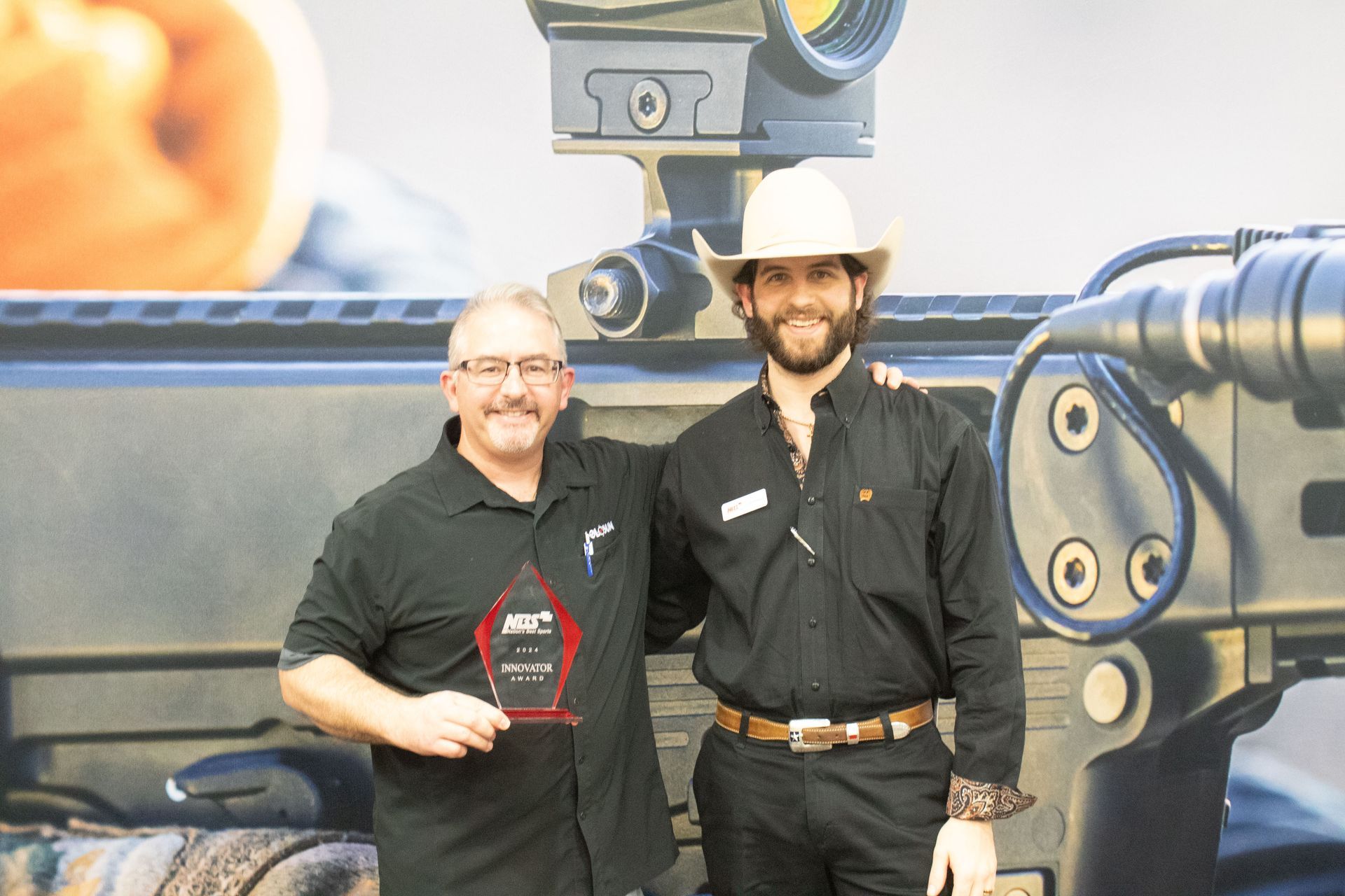 Two men standing in front of a large industrial machine, one in a white hat and black shirt, smiling.