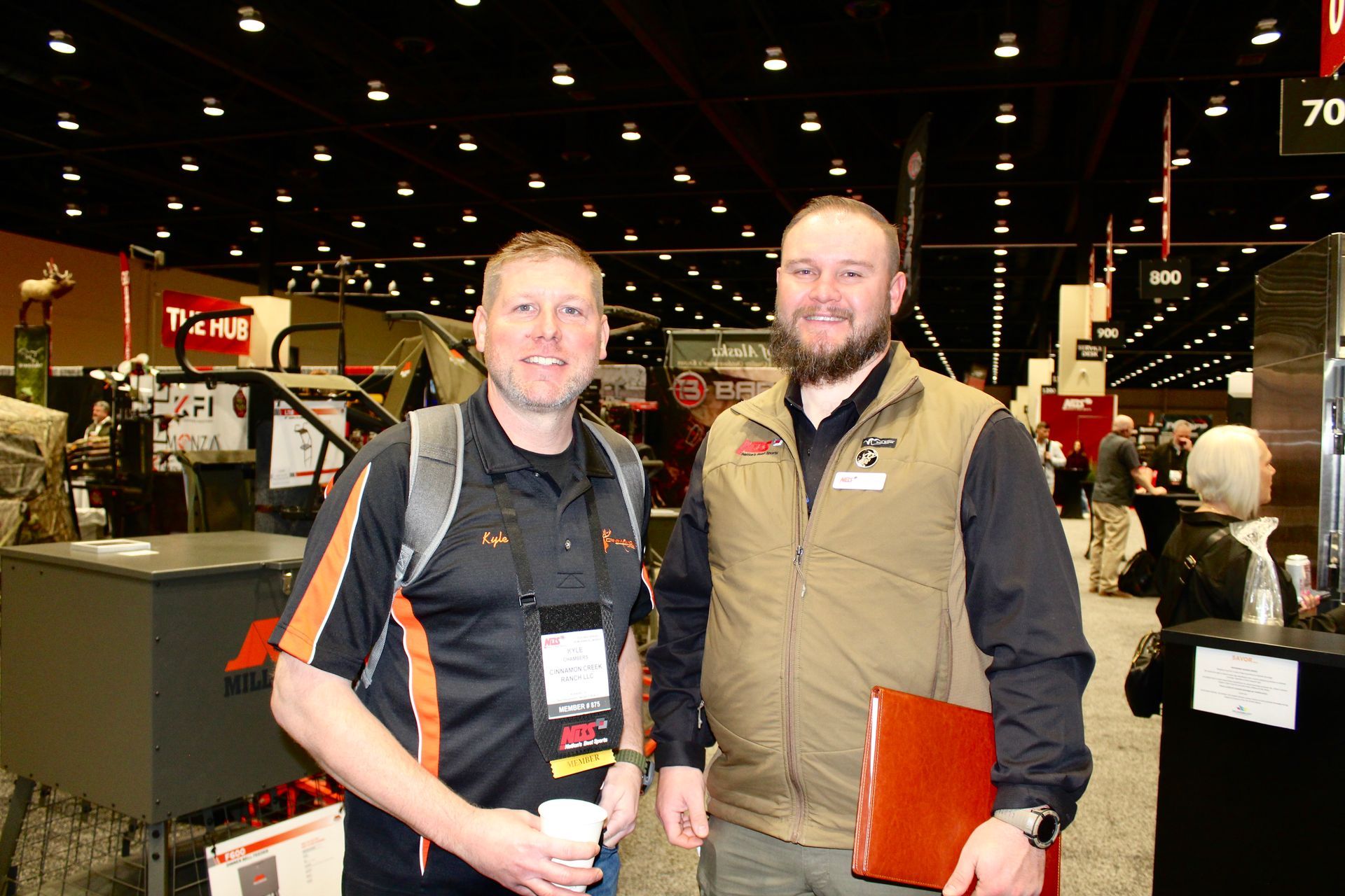 Two men standing at an indoor trade show, one holding a cup and the other an orange folder.