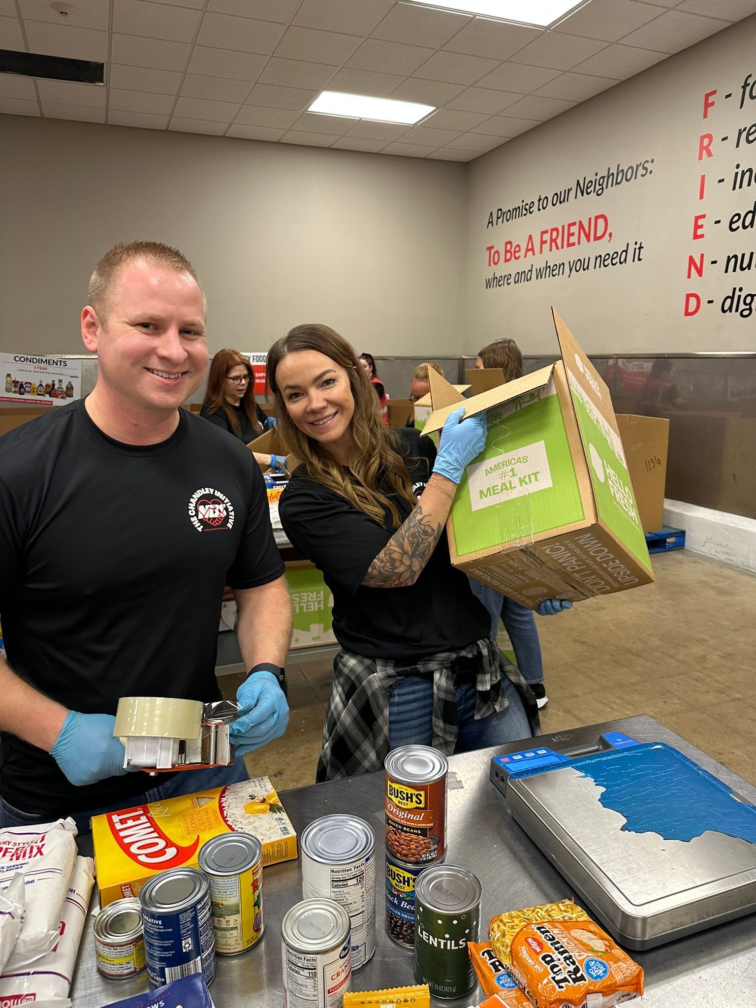 Two people packing food boxes in a classroom with cans and trays on the table