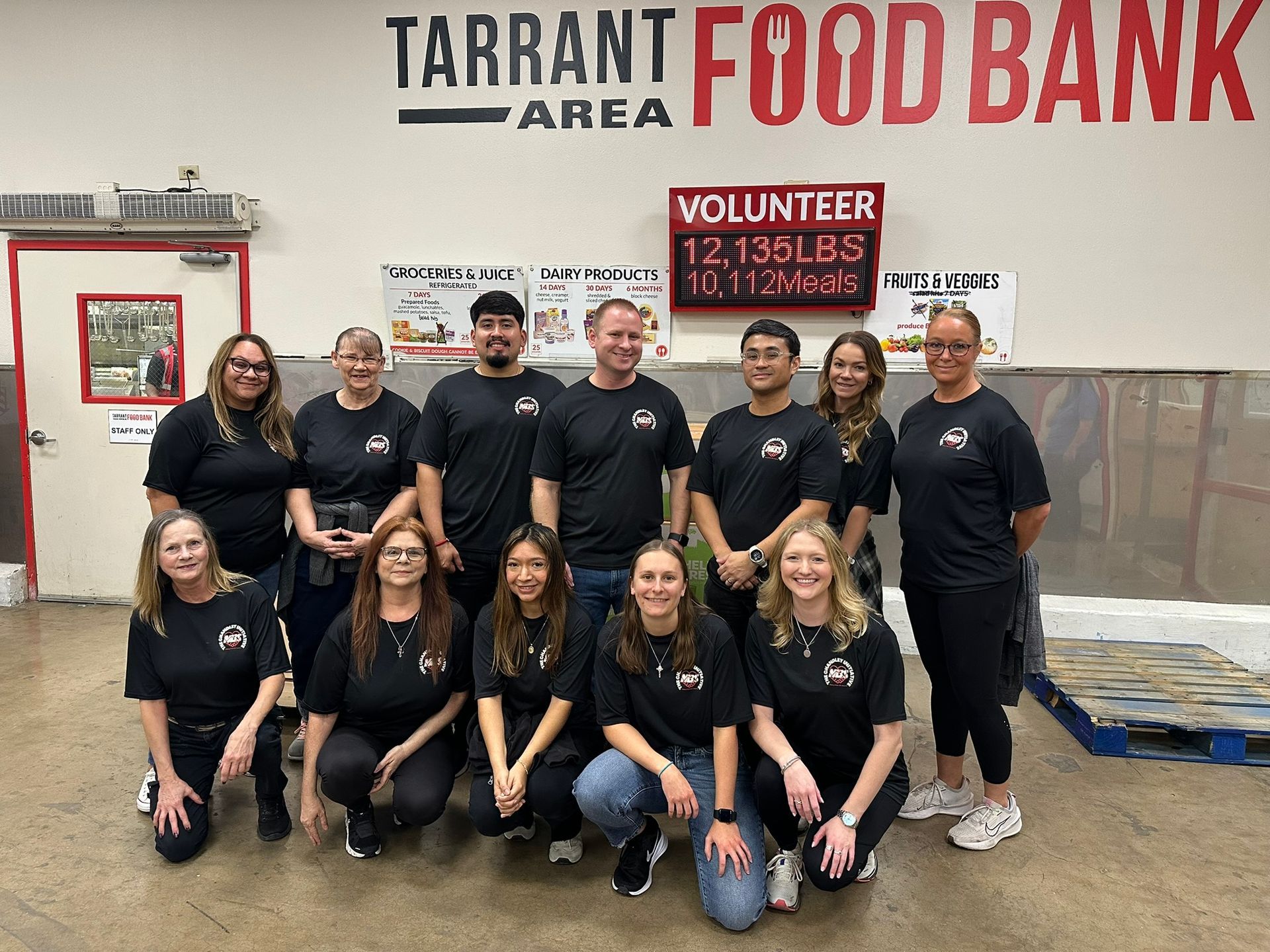 Volunteers posing inside Tarrant Area Food Bank warehouse, with logo and stacked pallets behind them.