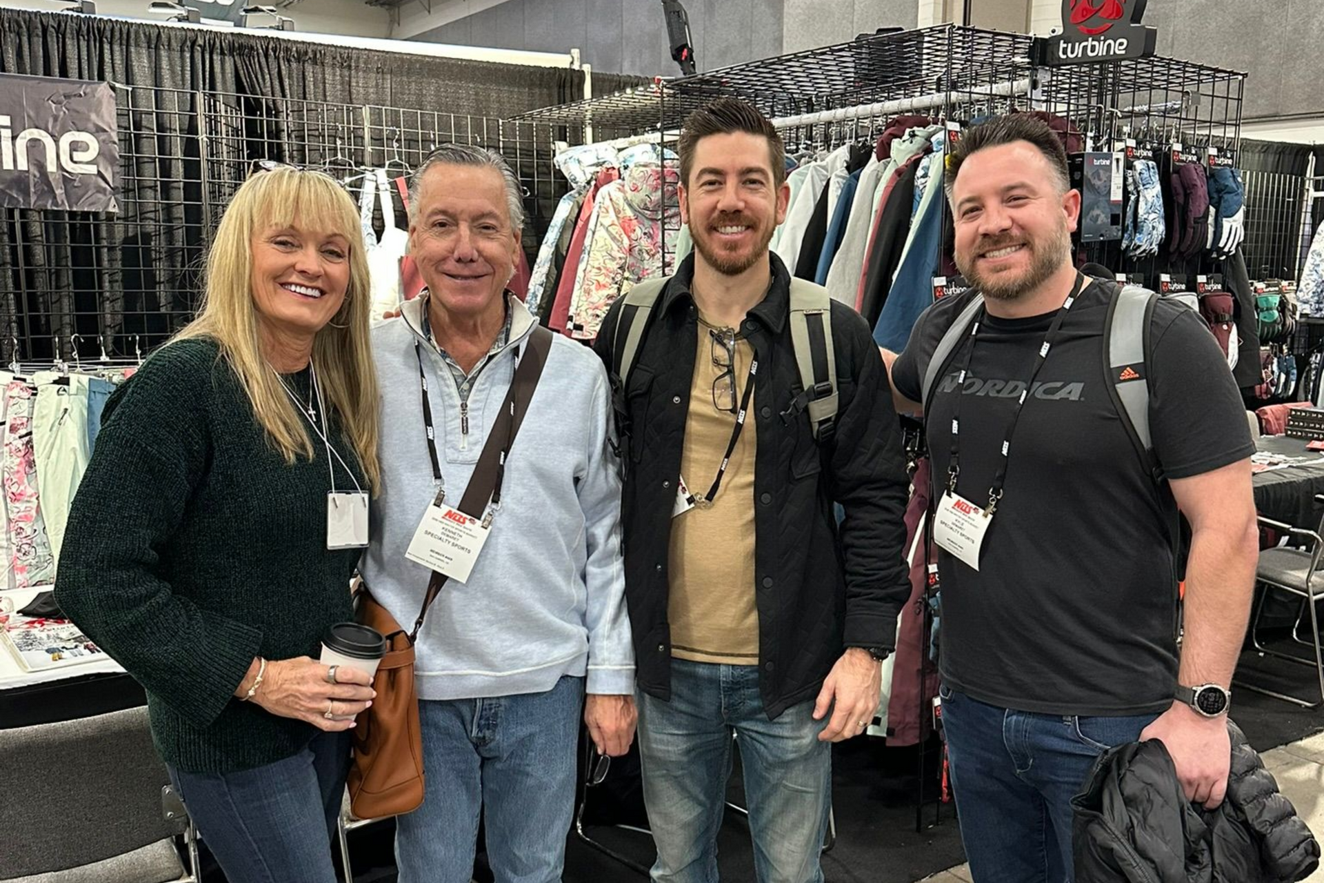 Four people standing together at an indoor market booth with hanging gear displays, smiling at the camera.