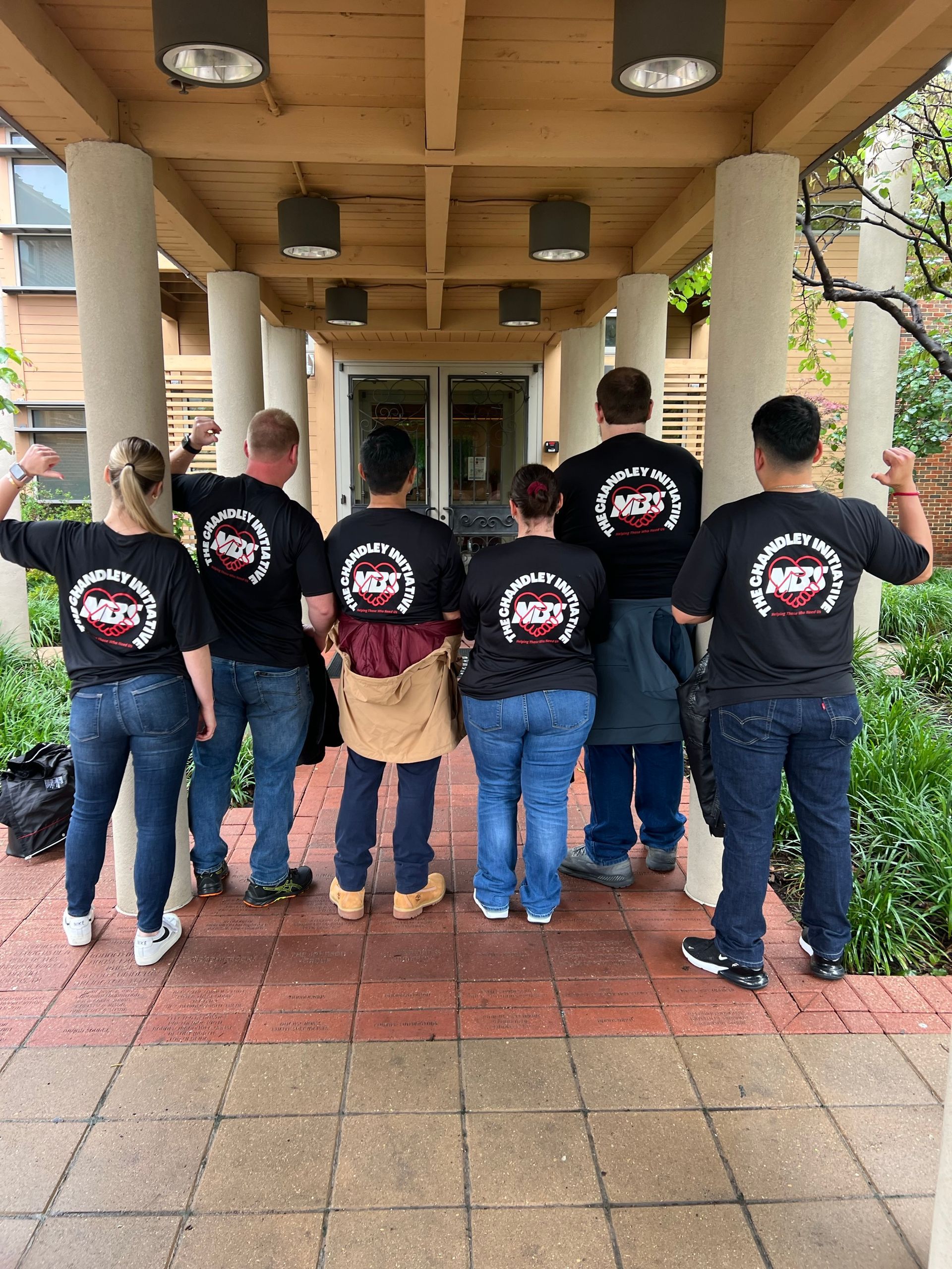 Seven people in black shirts stand under a covered walkway, seen from behind, with arms around each other.