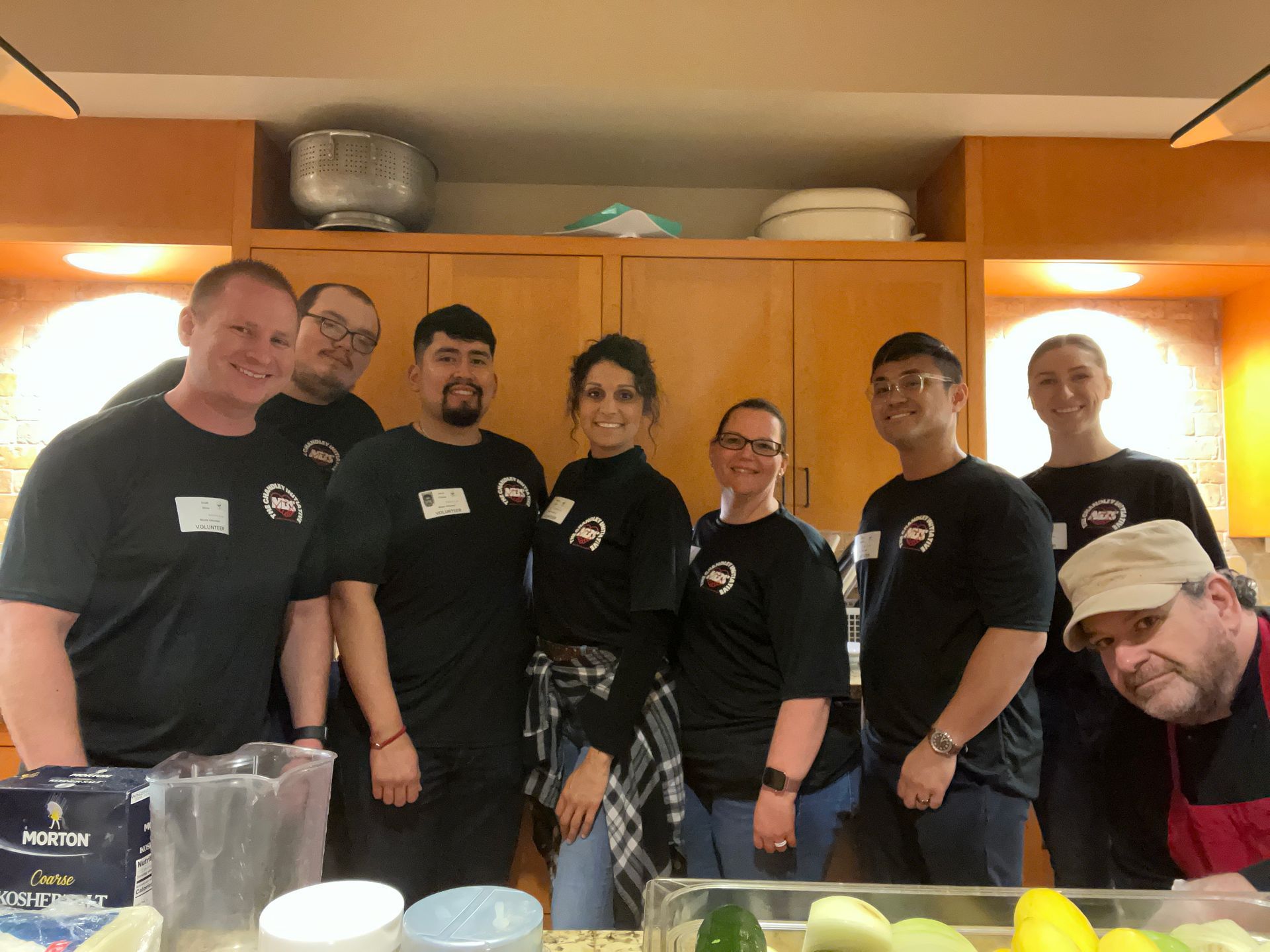 Group of people in black shirts posing together in a kitchen with wooden cabinets and warm lighting