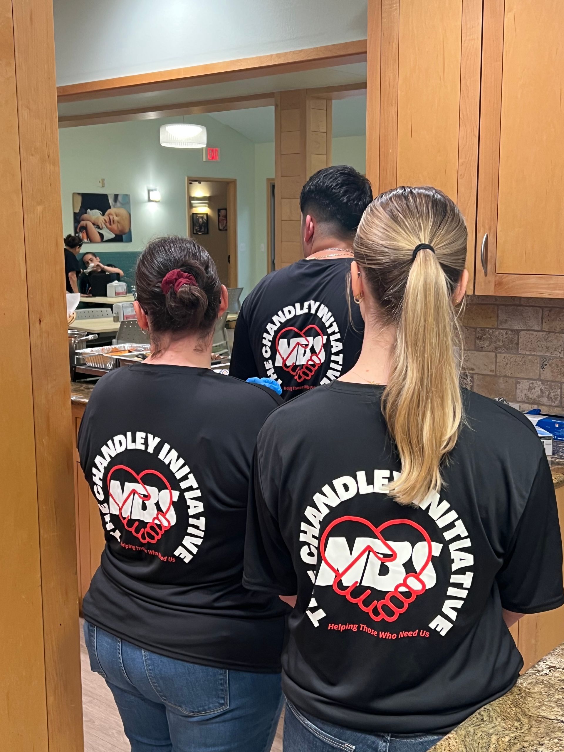 Three people in black matching shirts stand in a kitchen facing a mirror.