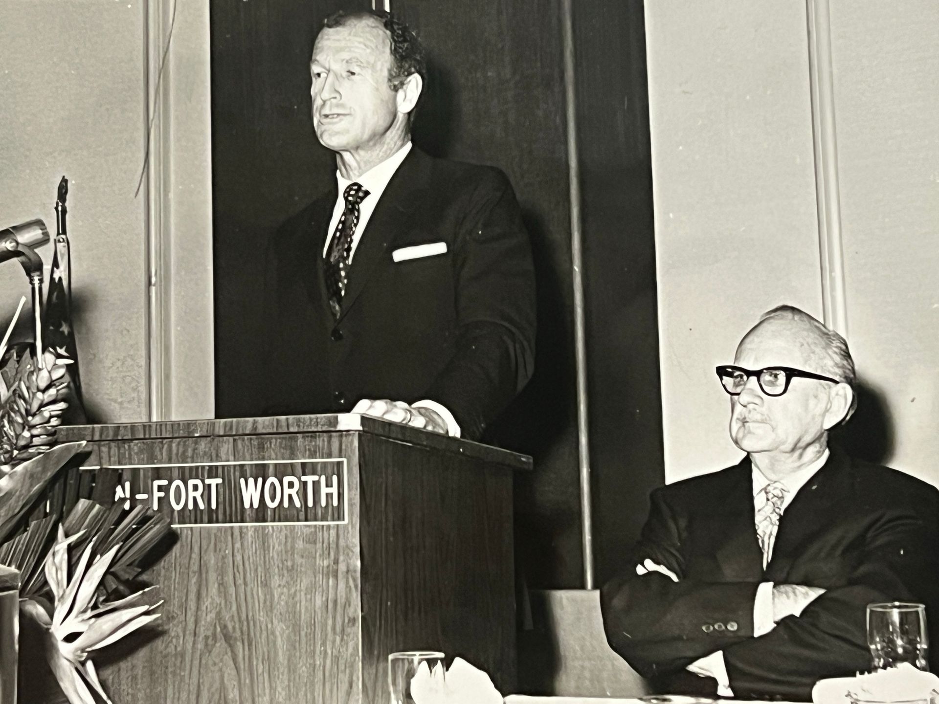 Man speaking at a podium in Fort Worth while another man sits beside him listening