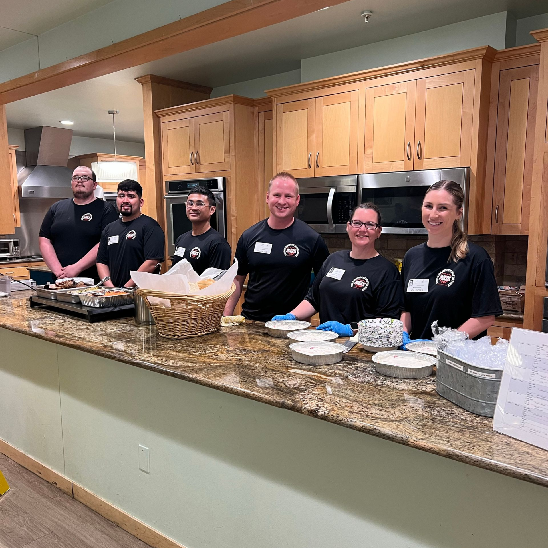 Five smiling volunteers in black shirts behind a kitchen counter with trays and bowls in a bright room