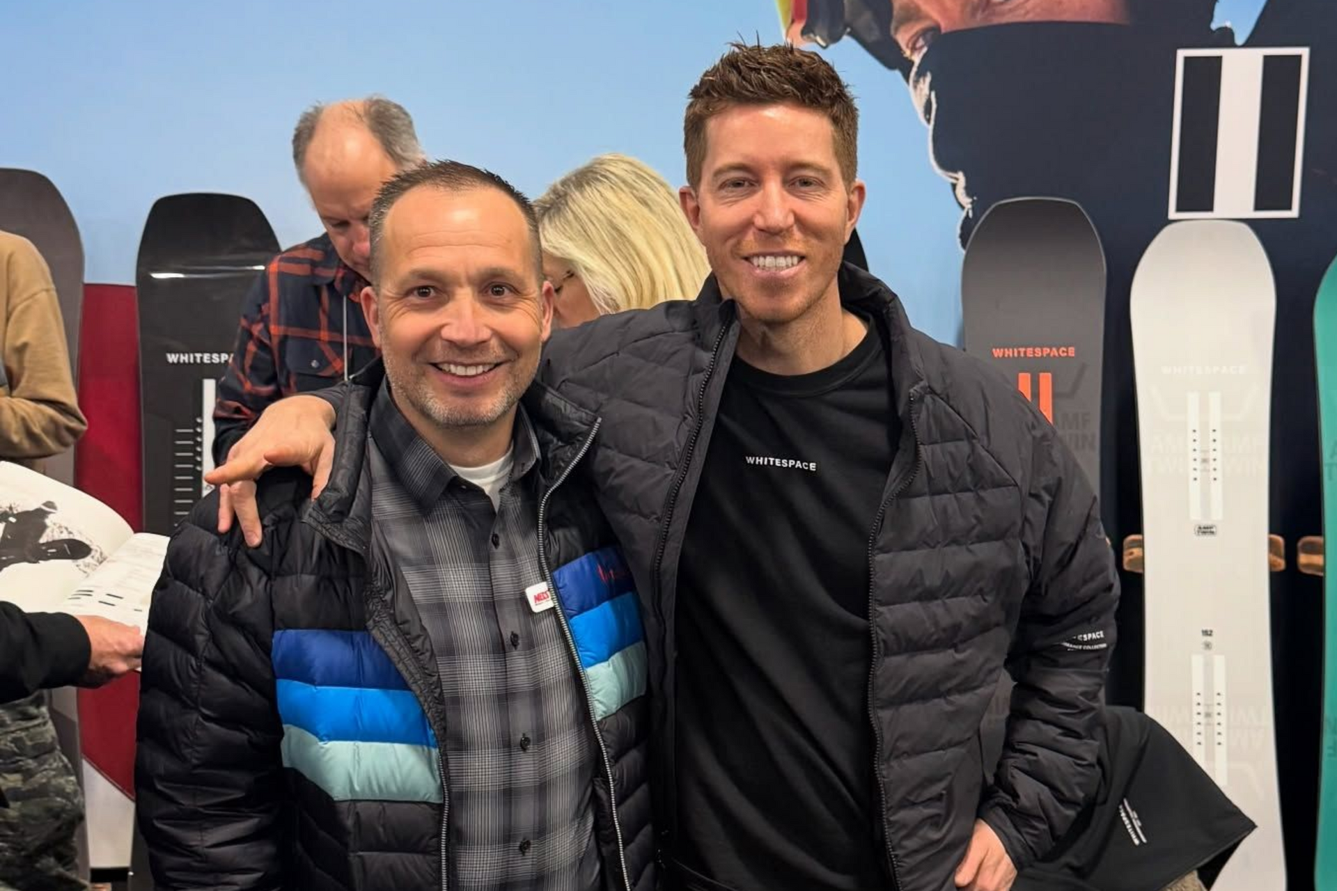 Two men smiling at an indoor event, one in a black jacket and one in a black-and-blue vest, with snowboards behind them
