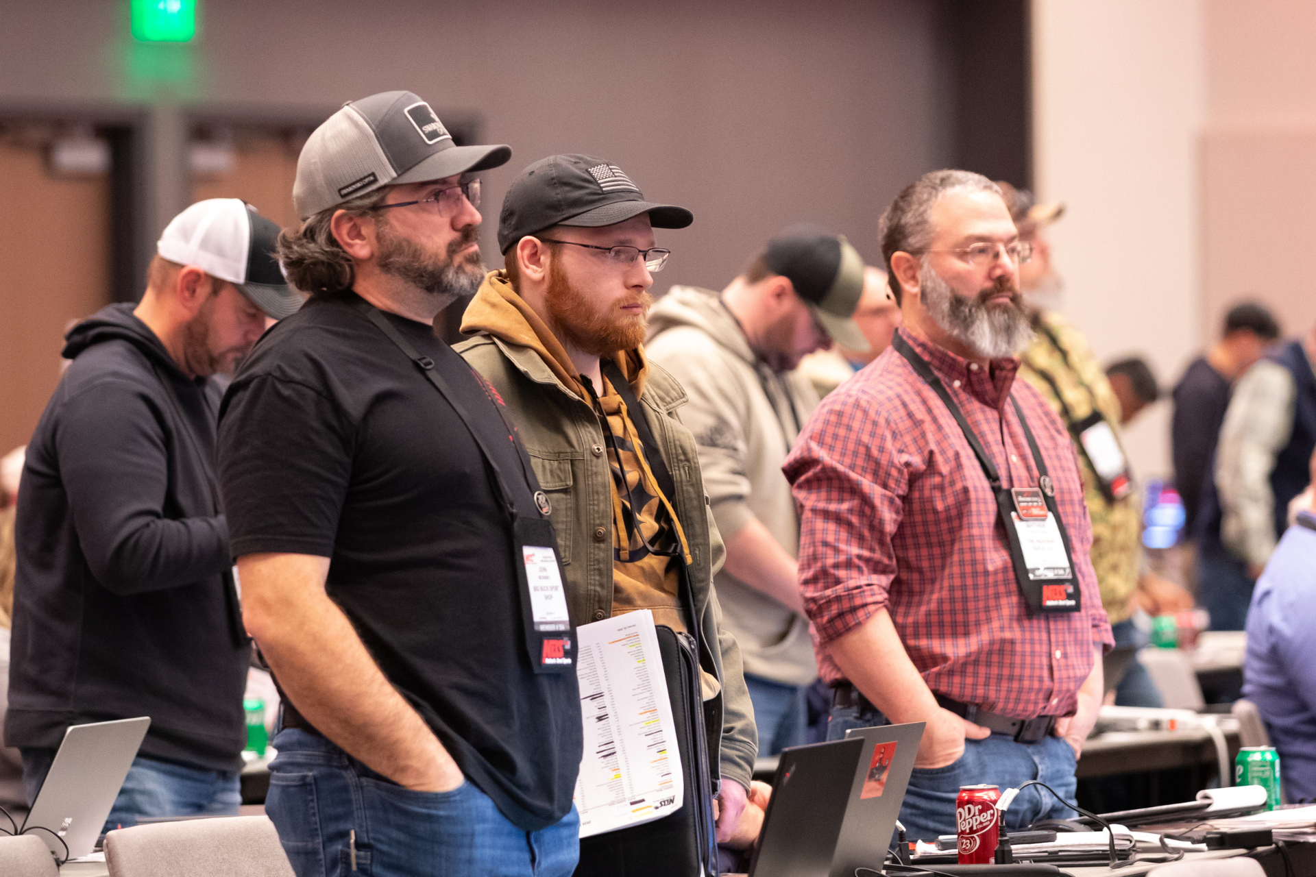 Attendees standing at a conference table, wearing casual clothes and badges, listening attentively