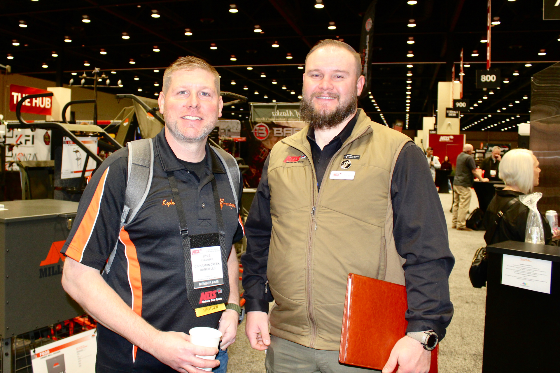Two smiling men at an indoor trade show, one holding a coffee cup and the other an orange folder.