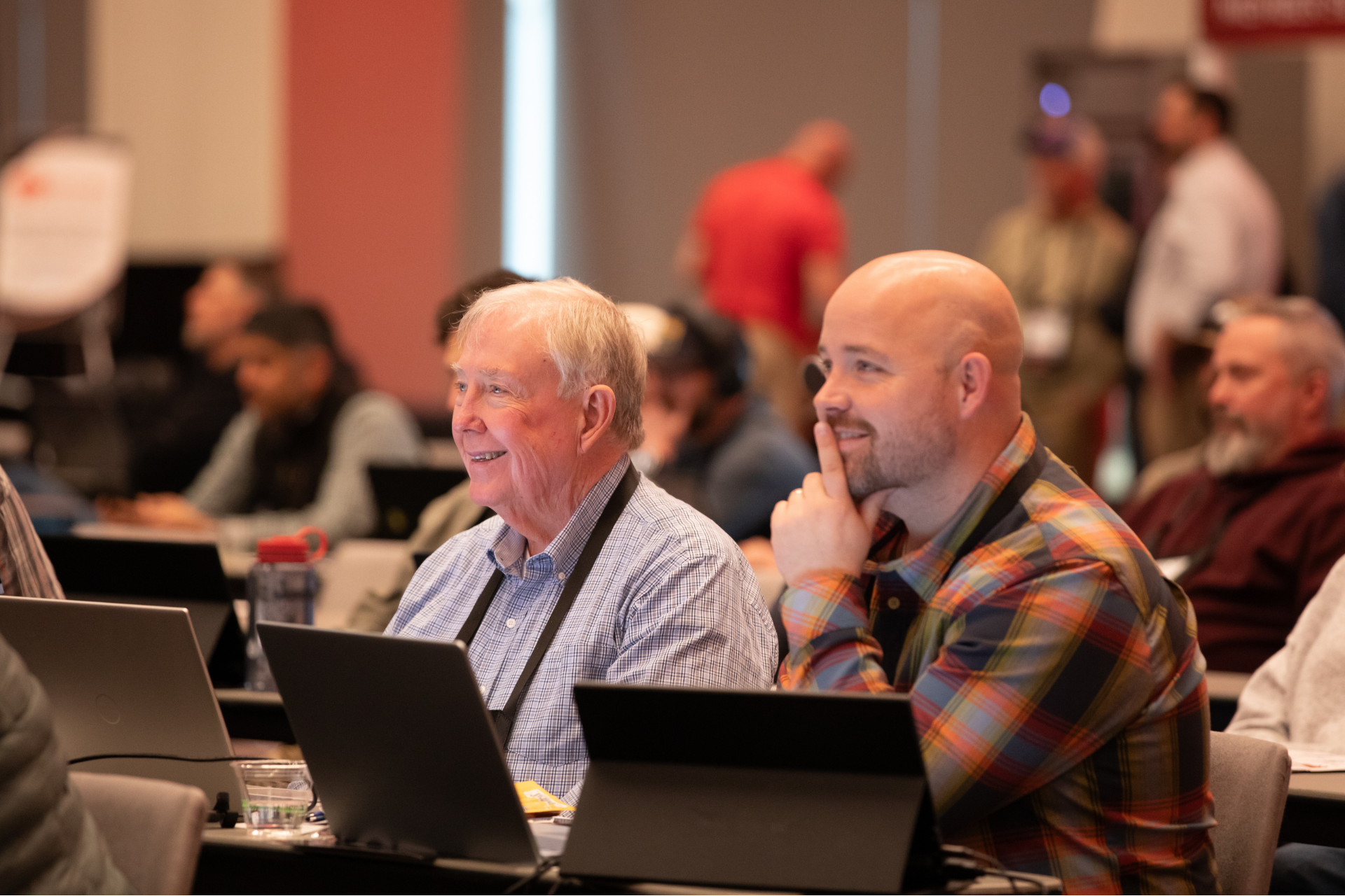 Conference attendees working on laptops at a table, with one man resting his chin on his hand.