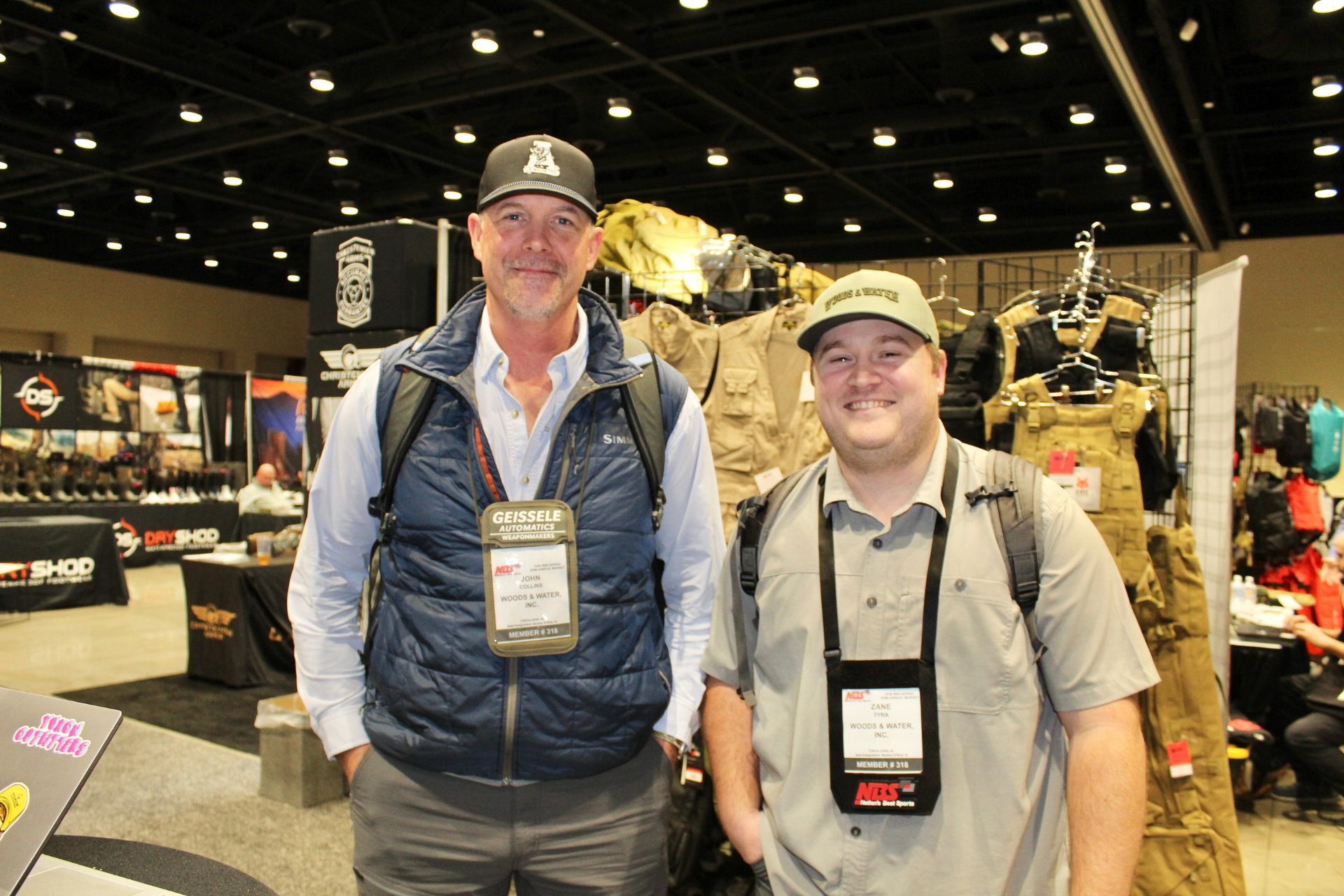 Two men smiling at an indoor market booth with hunting gear displays behind them