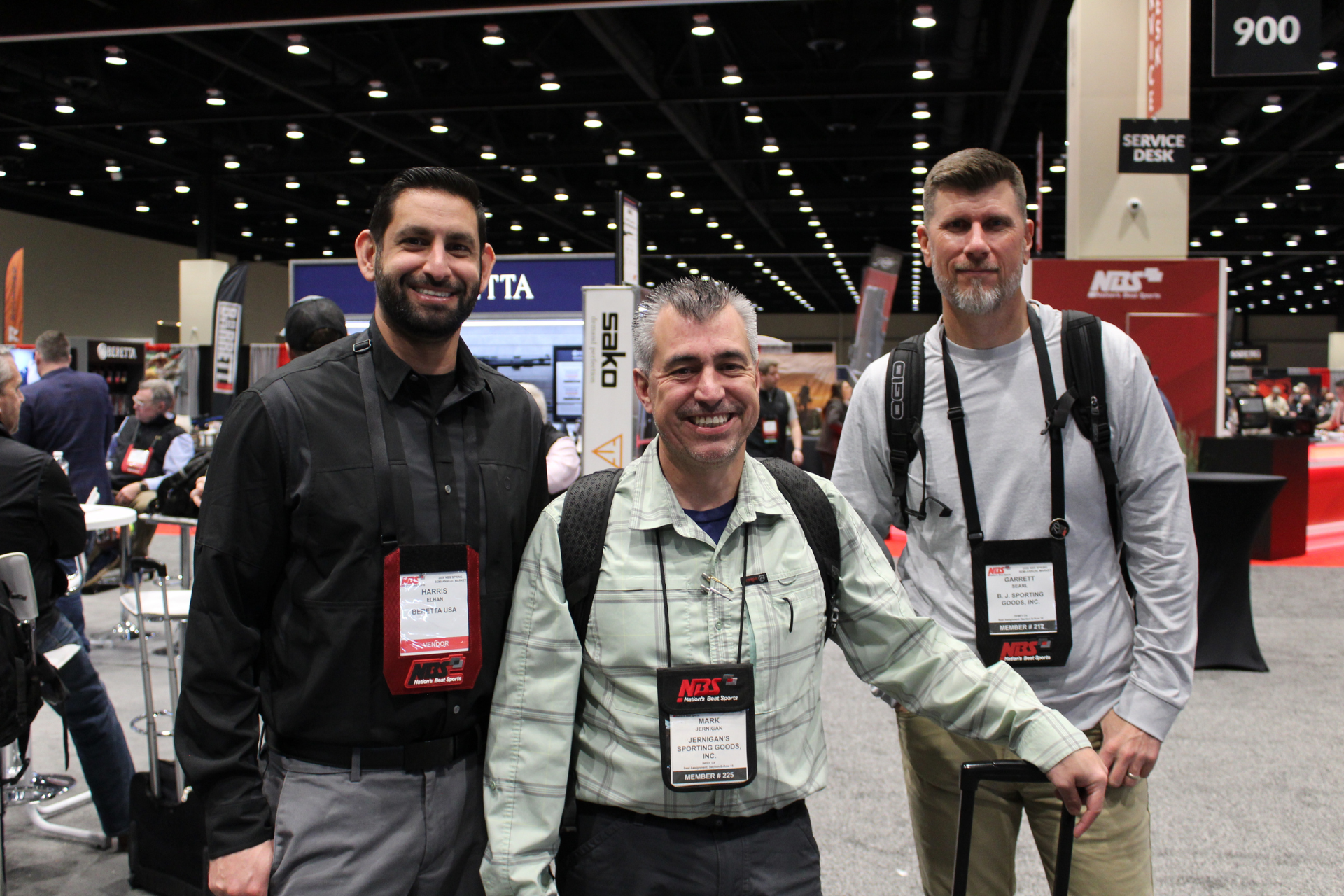 Three men posing at a trade show booth, wearing conference badges and backpacks