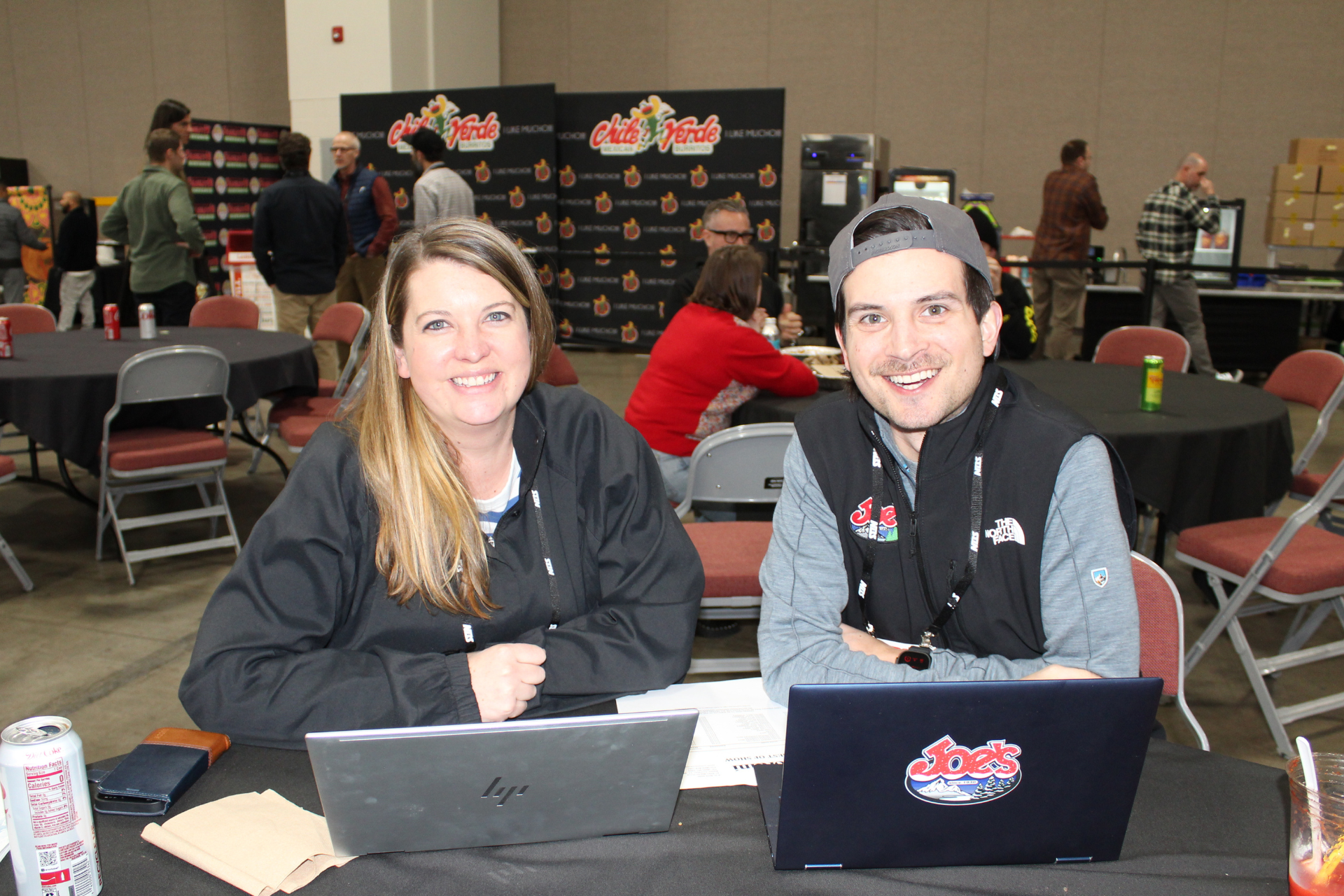Two people at a table with laptops at an indoor event, smiling toward the camera.